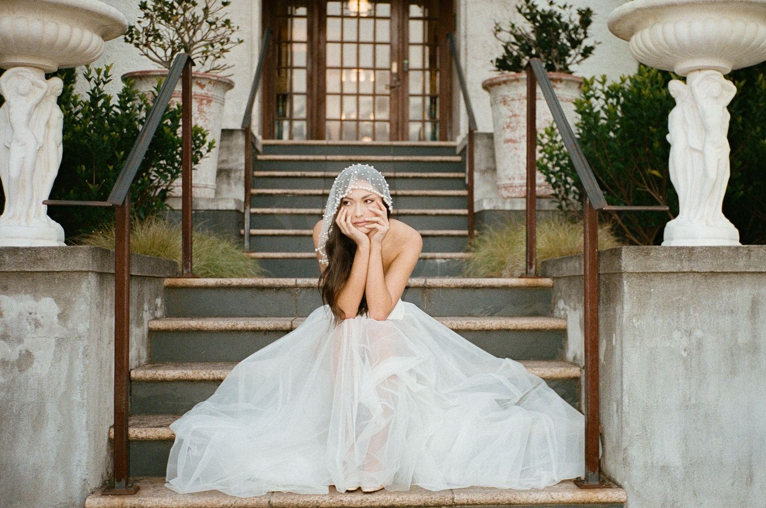 Bride in a flowing tulle gown and crystal crown sits gracefully on stone steps of an elegant venue.