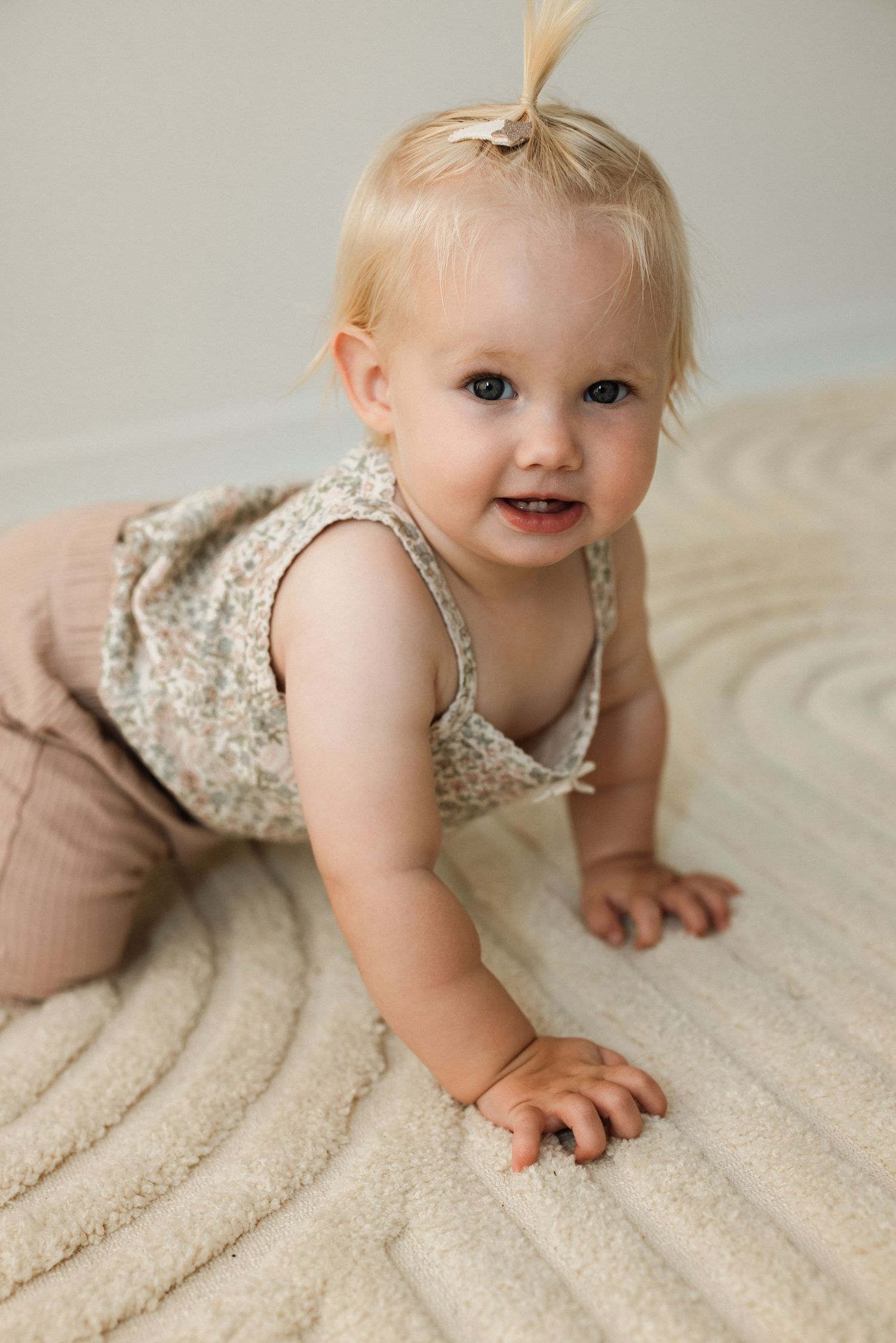 Baby in floral dress and tan pants crawling on textured beige blanket.