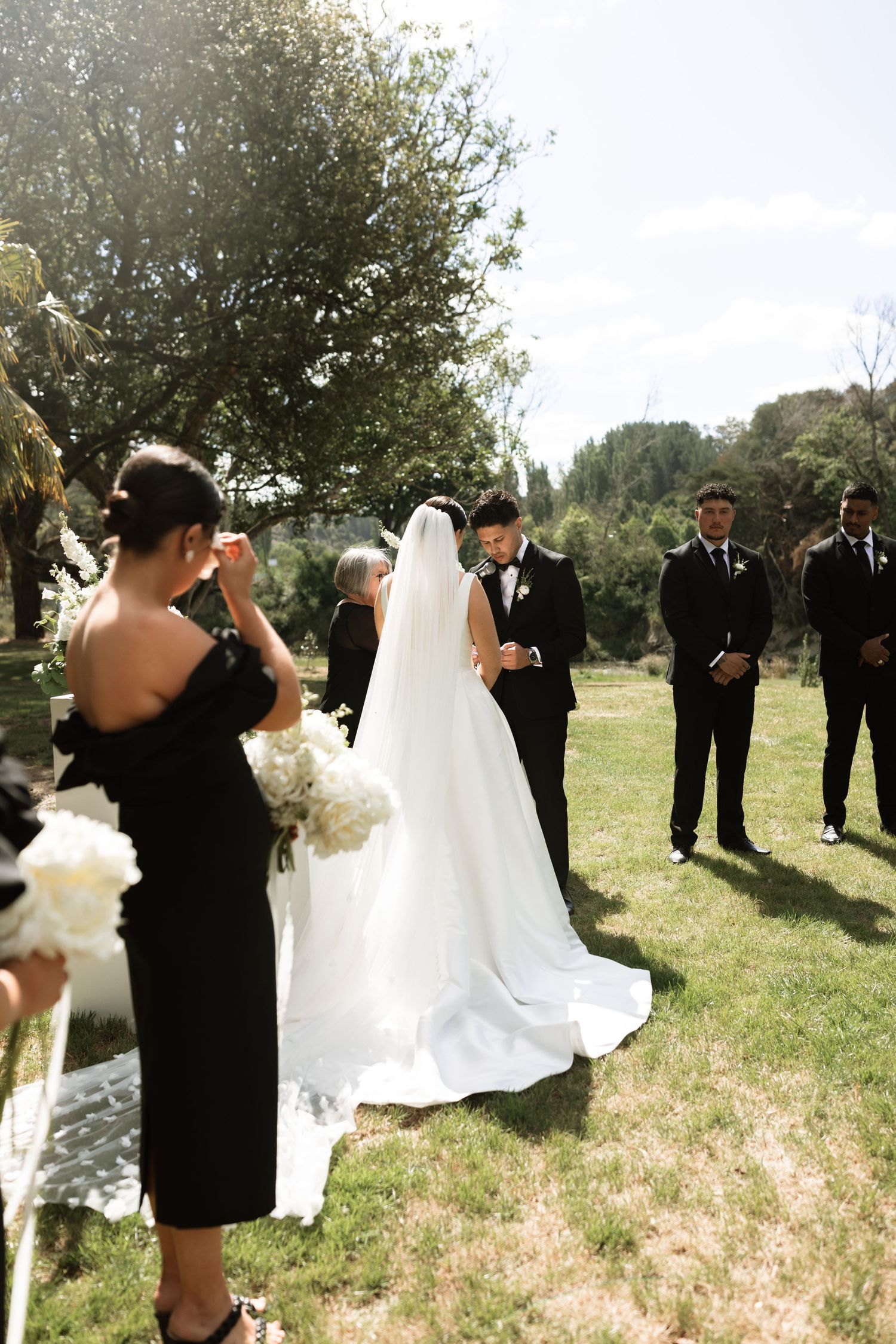 Wedding party celebrates outdoors with flowing white dress and formal attire.