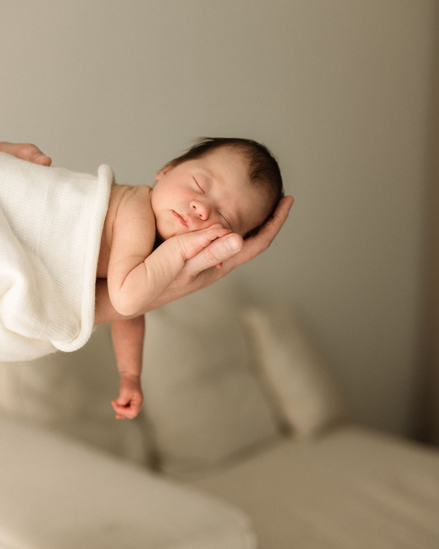 Newborn baby sleeping peacefully on a parent's hand, wrapped in a white blanket, in a soft neutral studio setting.