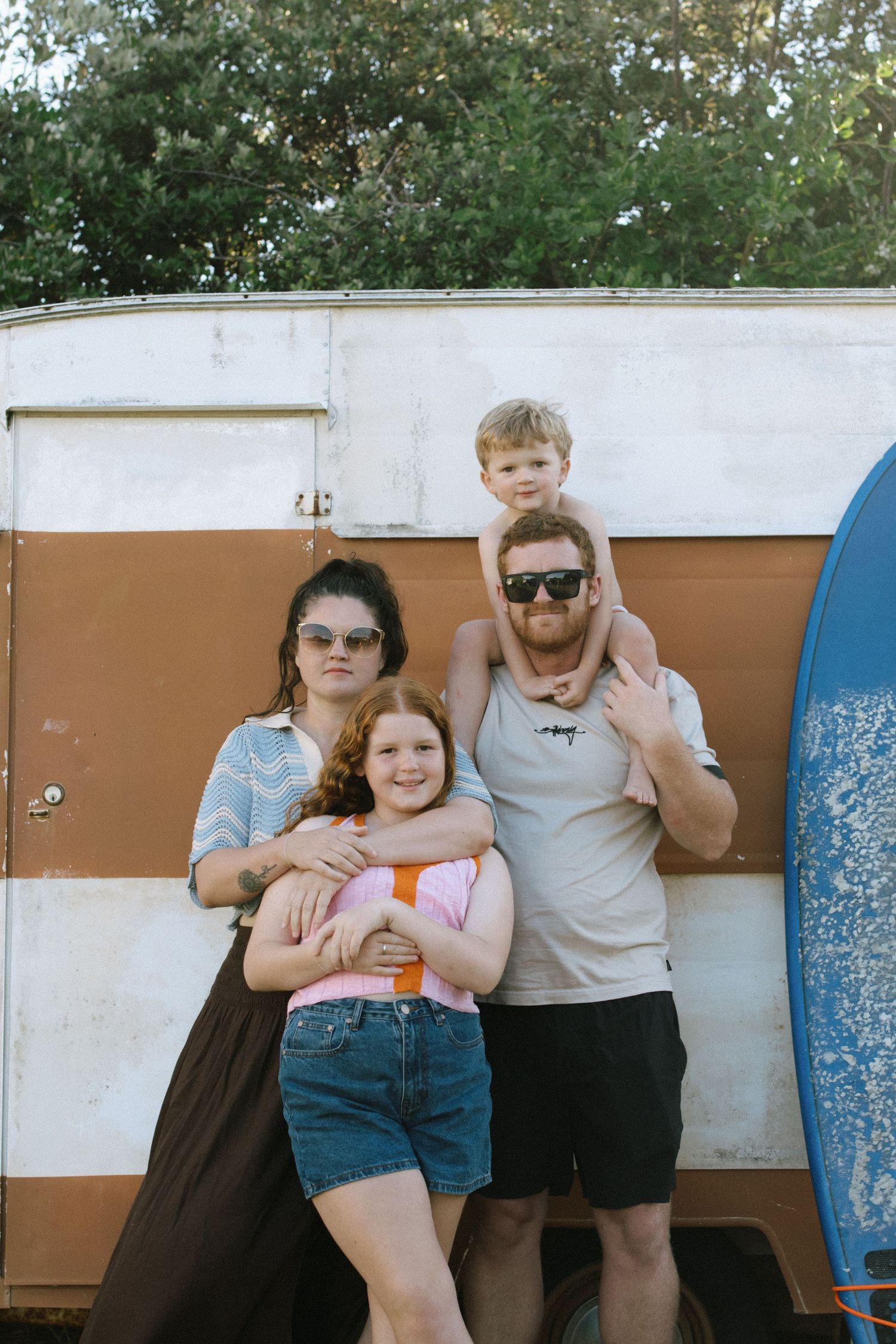 Family of four posing in front of a vintage brown and white caravan with a surfboard, trees in background.