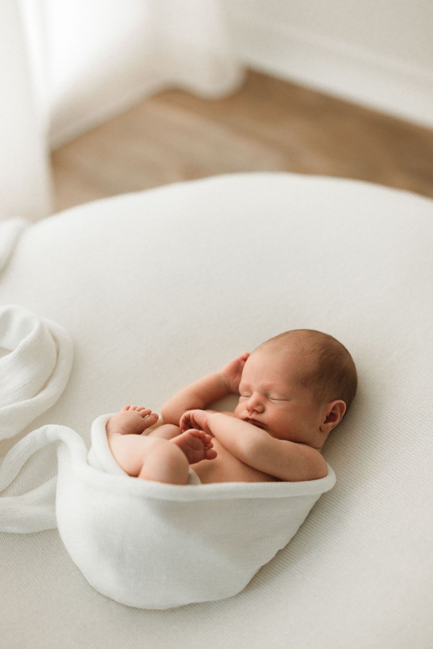 Sleeping newborn baby wrapped in white blanket on light surface with minimalist styling.