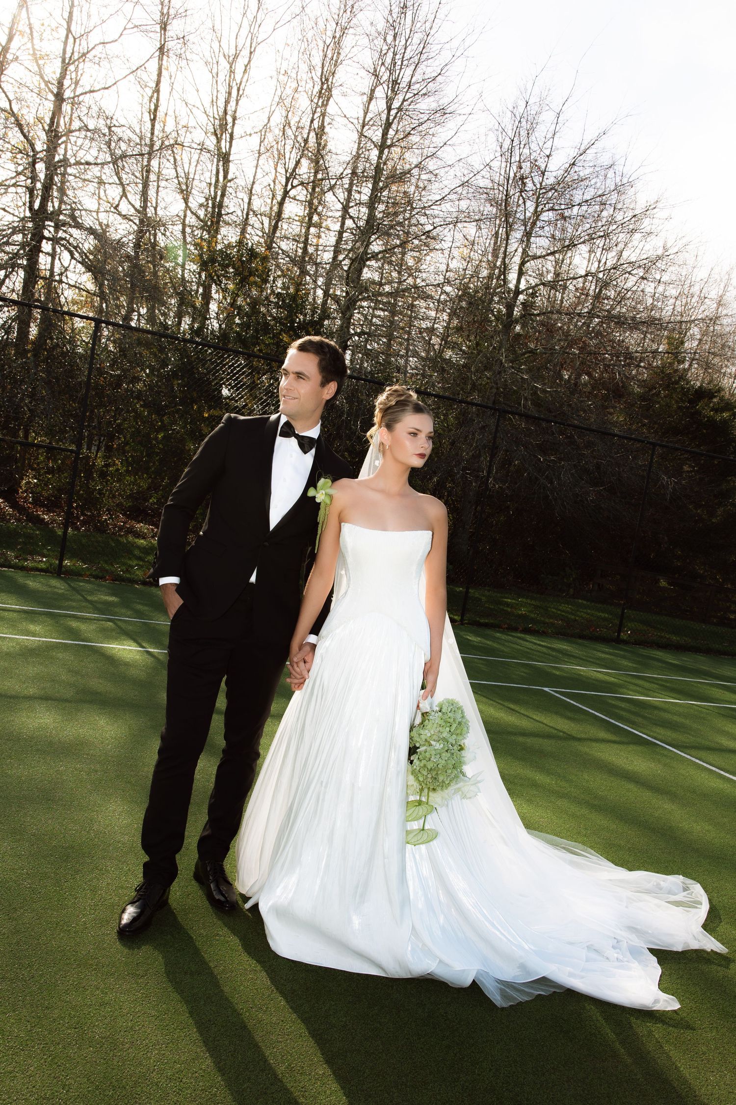 Newlyweds pose together on a sunlit tennis court with flowing wedding dress.
