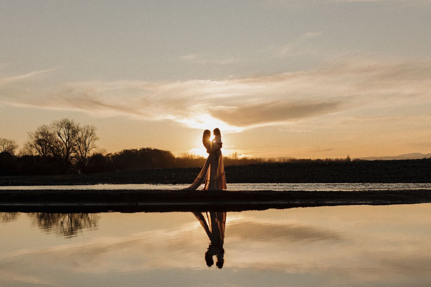 Silhouettes of two figures on a beach during golden sunset reflected in calm water.