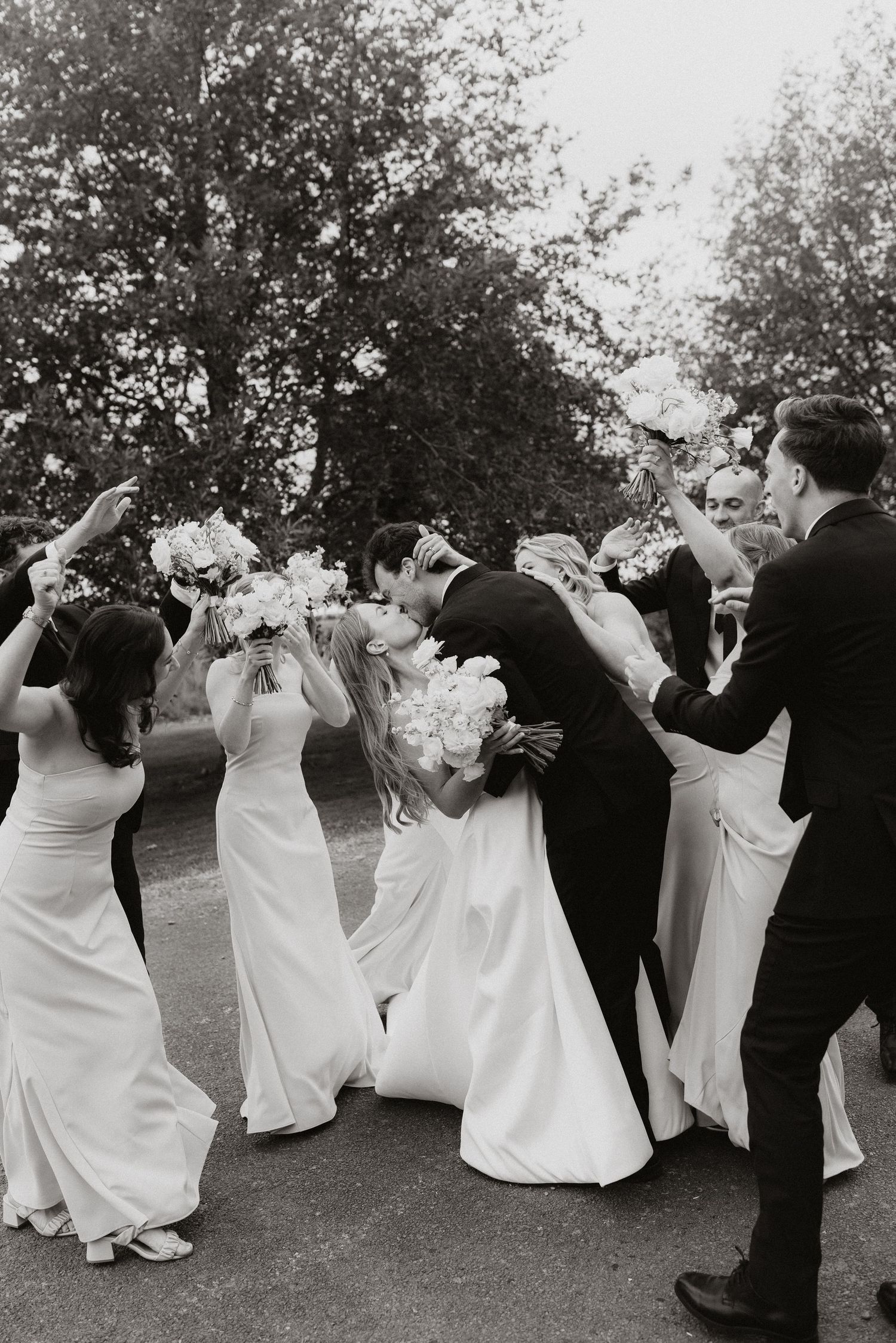 A wedding party in formal attire dips and dances together outdoors in a playful black and white photograph.