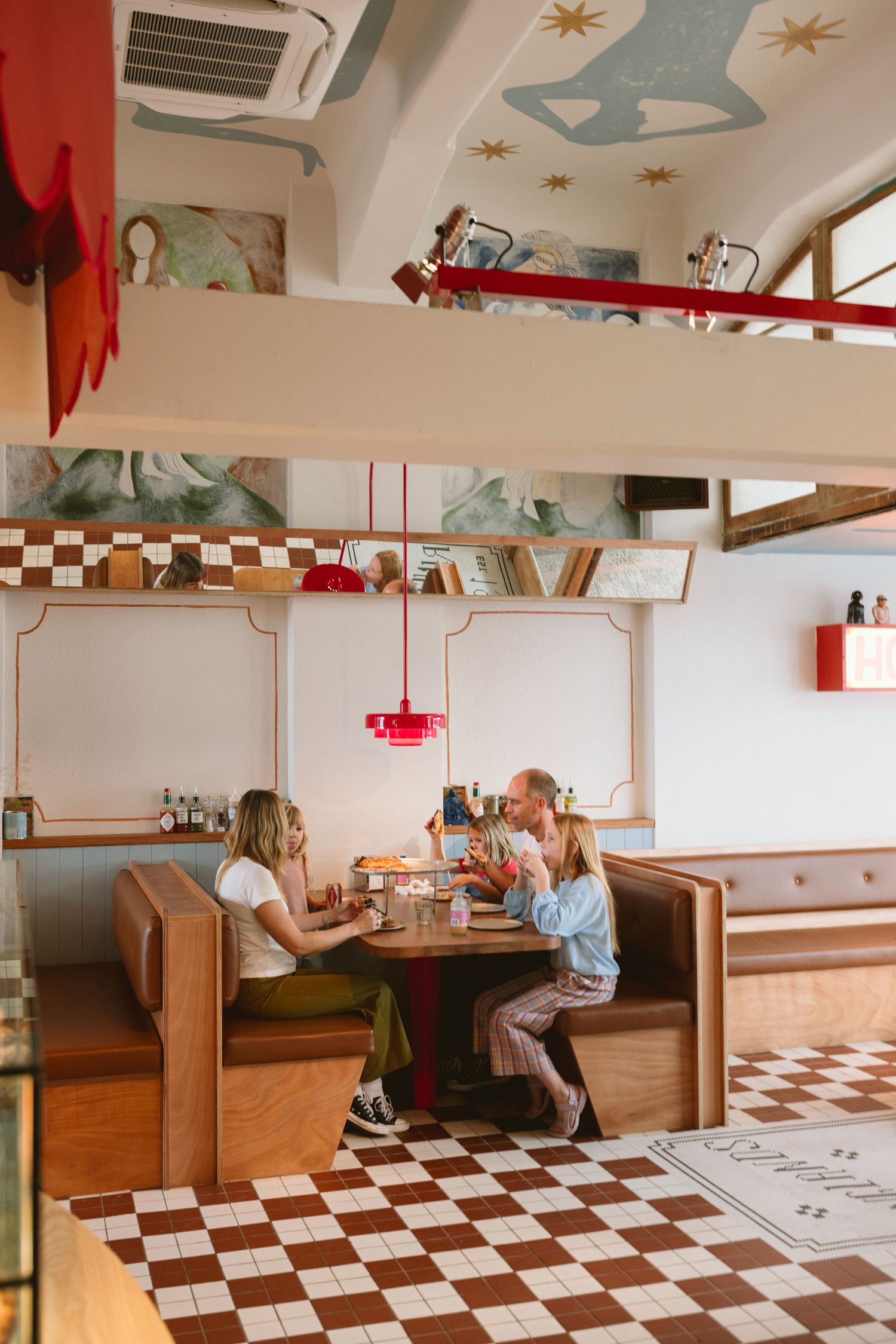 Three people dining in a retro diner booth with checkered floors and a red pendant light.