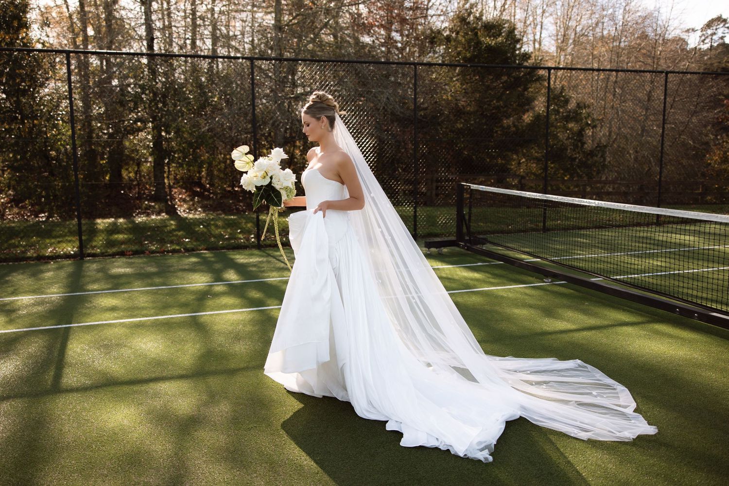A bride in a flowing white gown and veil stands on a tennis court holding a bouquet at sunset.
