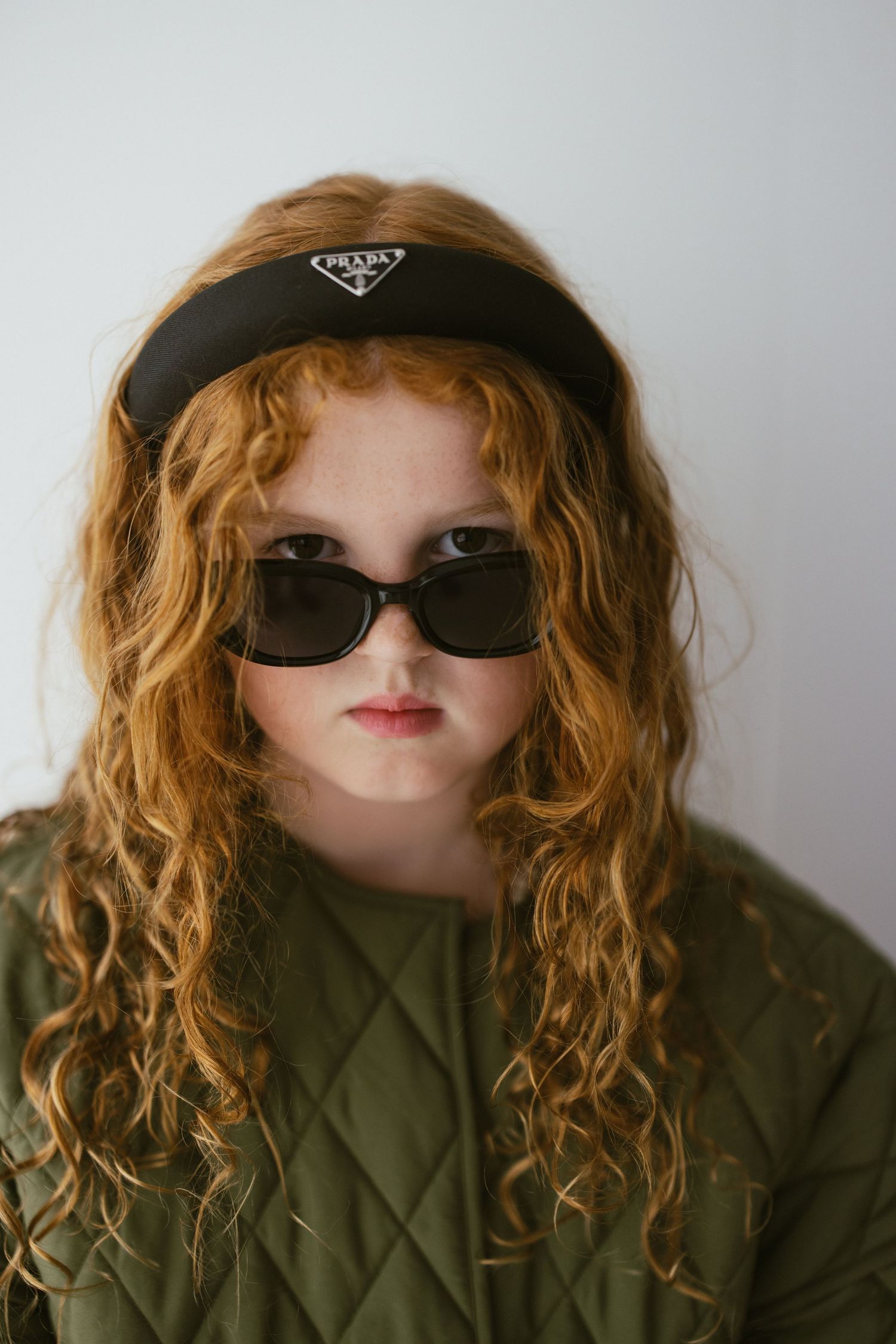 Young girl with curly red hair wearing Prada headband, dark sunglasses, and quilted green jacket.