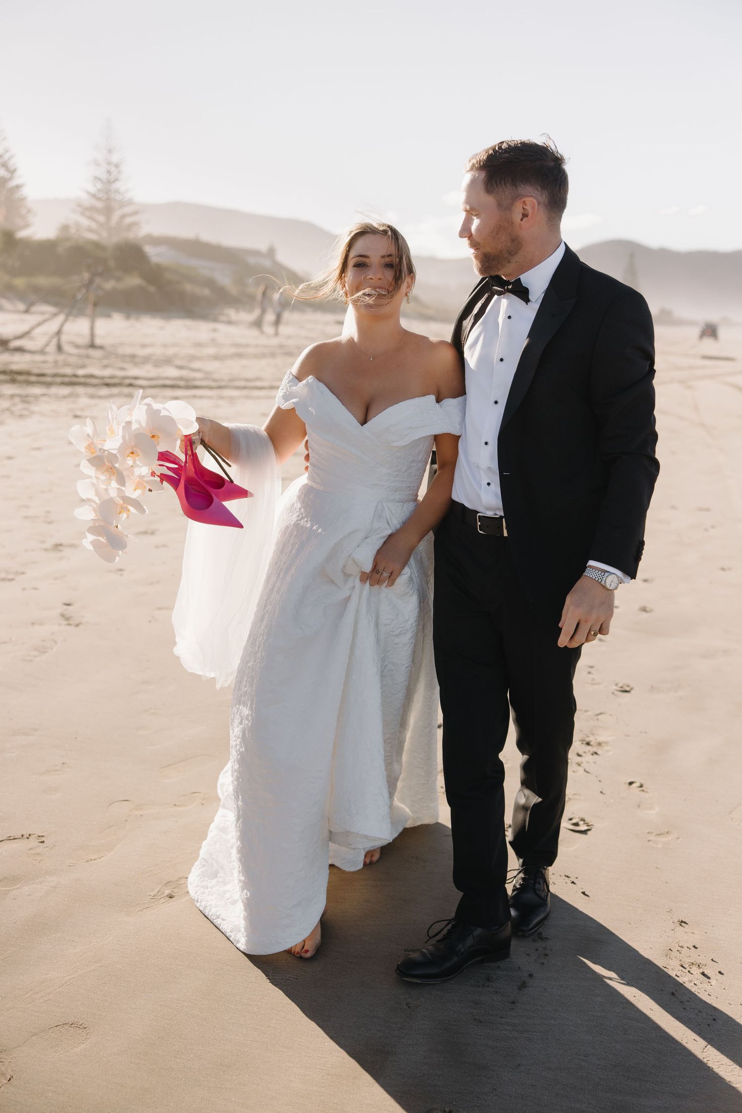 Couple in formal wedding attire walking along wooden boardwalk at beach during sunset.