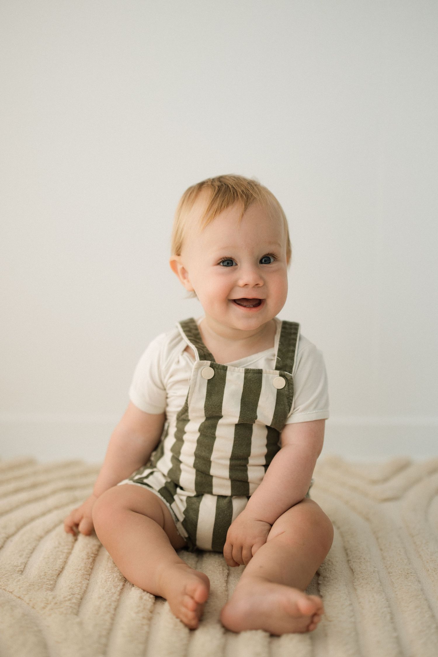 Baby in striped overalls sitting and laughing on cream colored blanket.