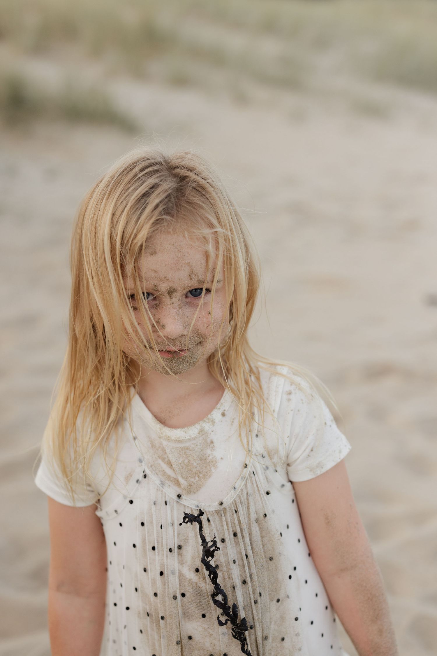 Person with blonde hair and white polka dot shirt sits looking downward in a natural sandy setting.