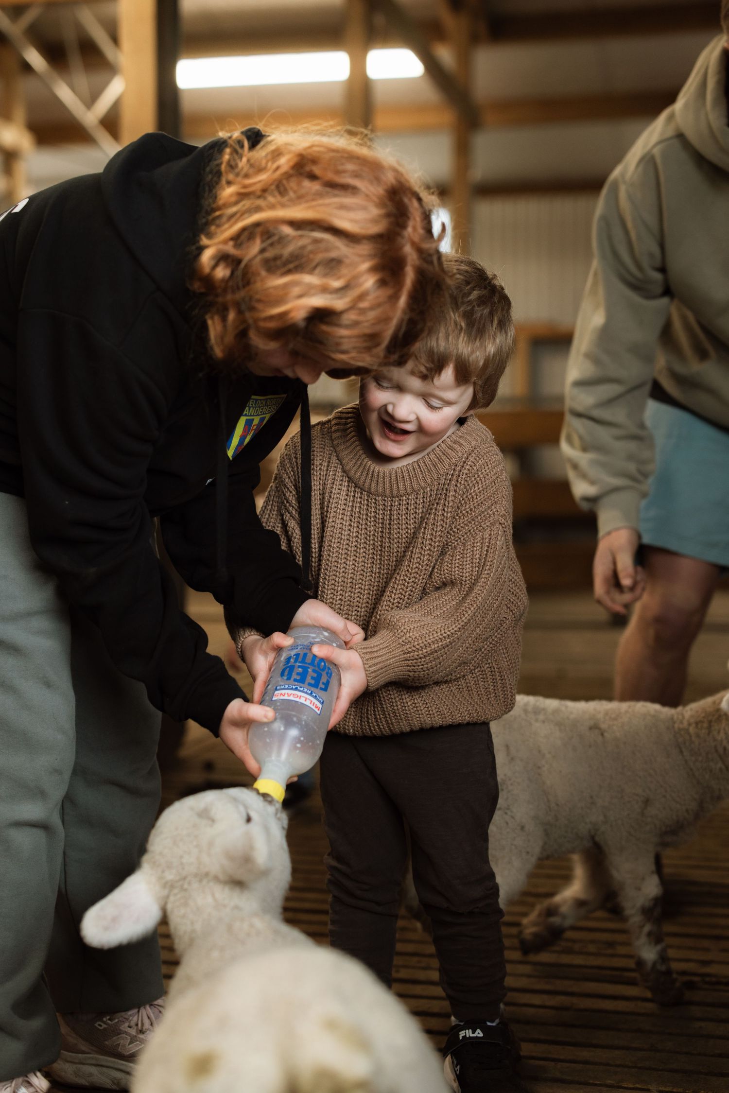 Child in brown sweater joyfully bottle-feeds a baby lamb while being assisted in what appears to be a barn setting.