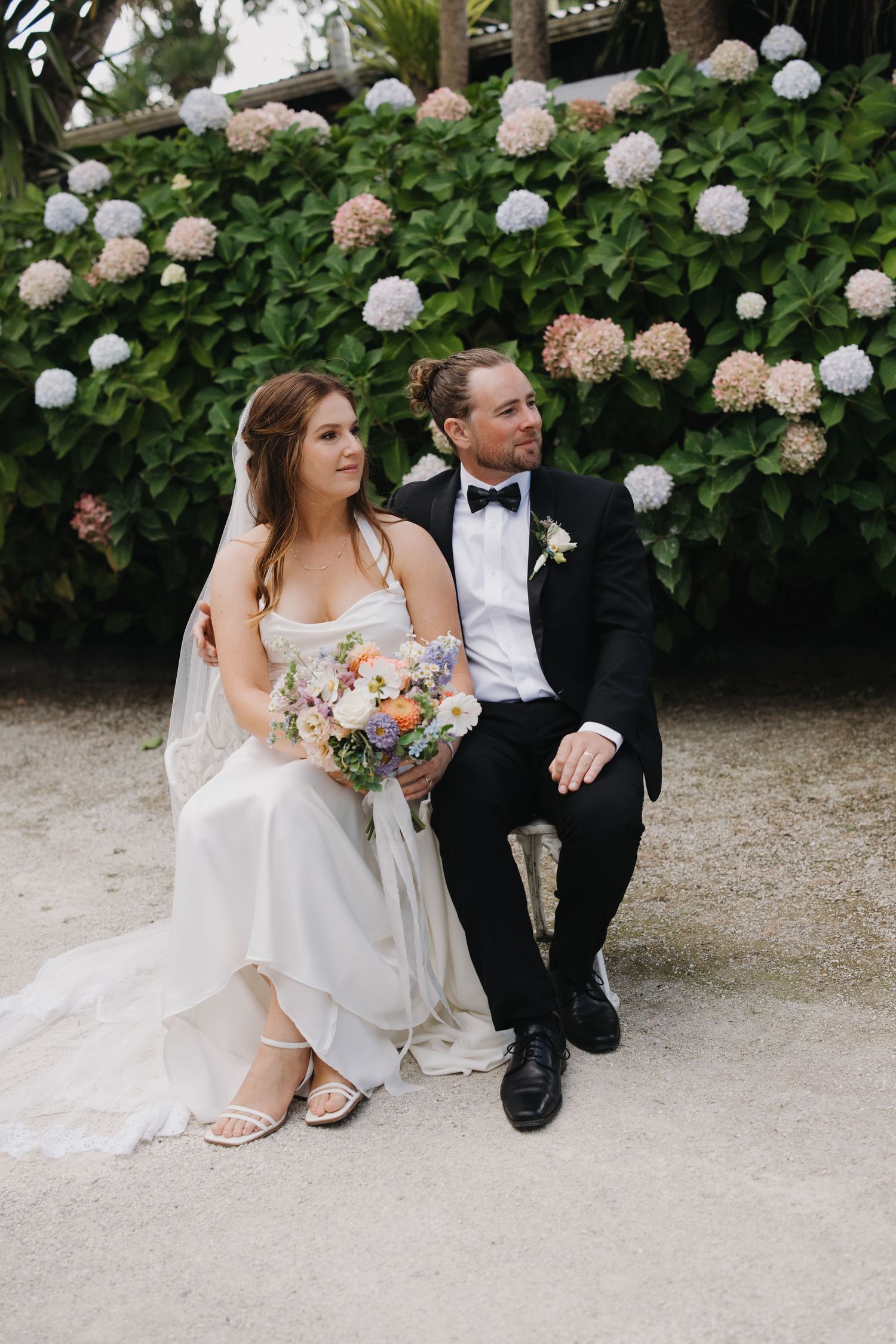 A wedding couple sits together on a bench surrounded by blooming white and pink hydrangea bushes in a garden setting.