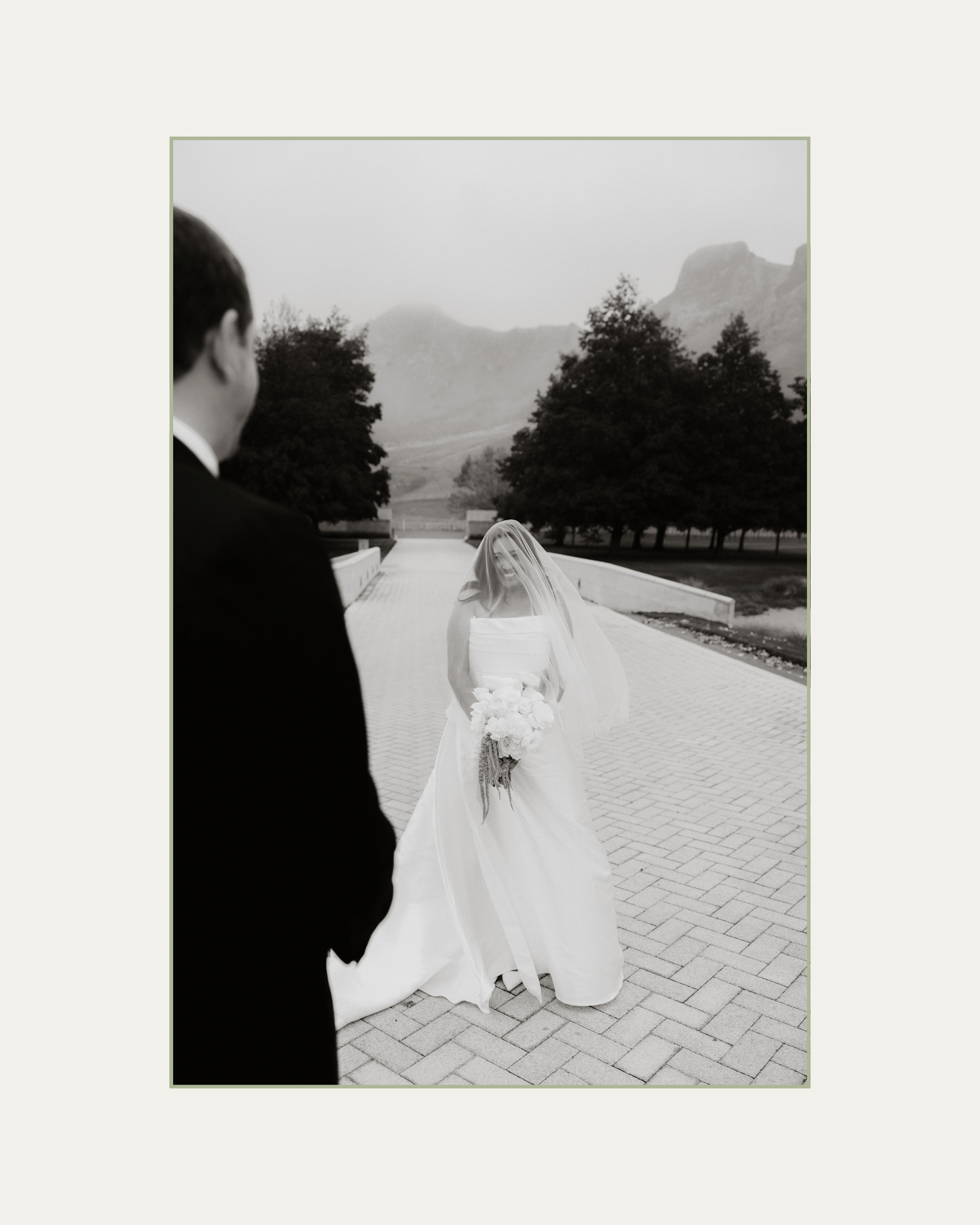 Black and white wedding photo of bride in white gown and veil holding bouquet, walking toward groom outdoors.