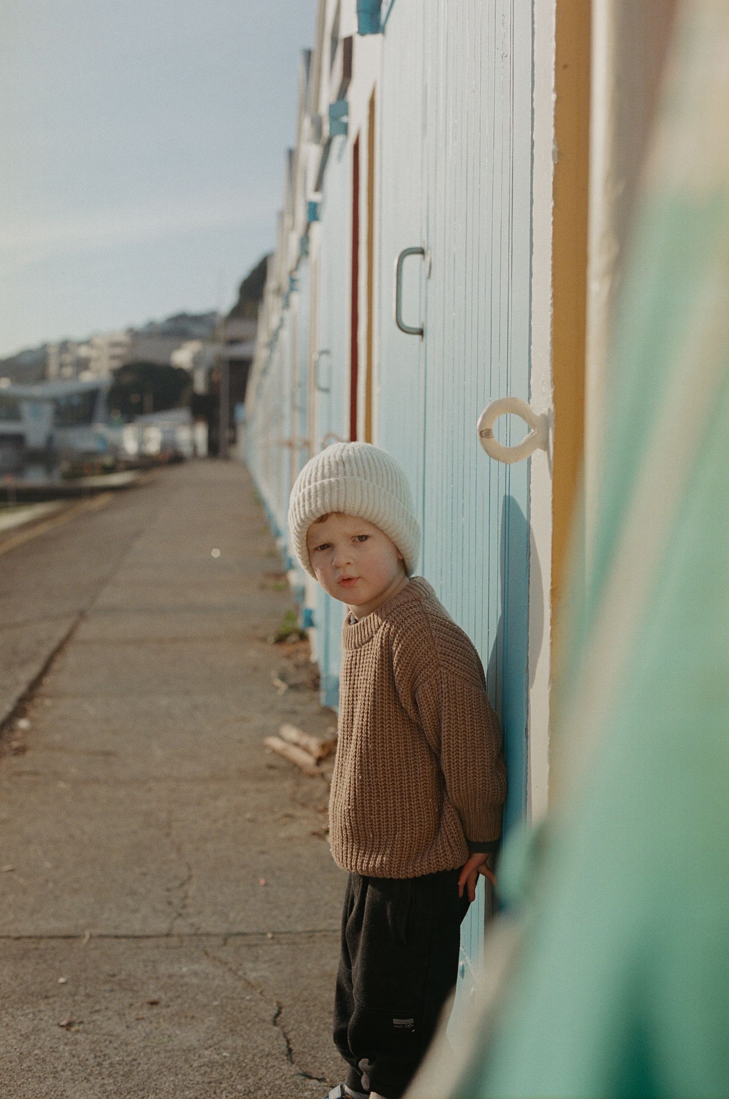 A person in a beige knit hat and brown sweater stands against a wall in a muted outdoor setting.