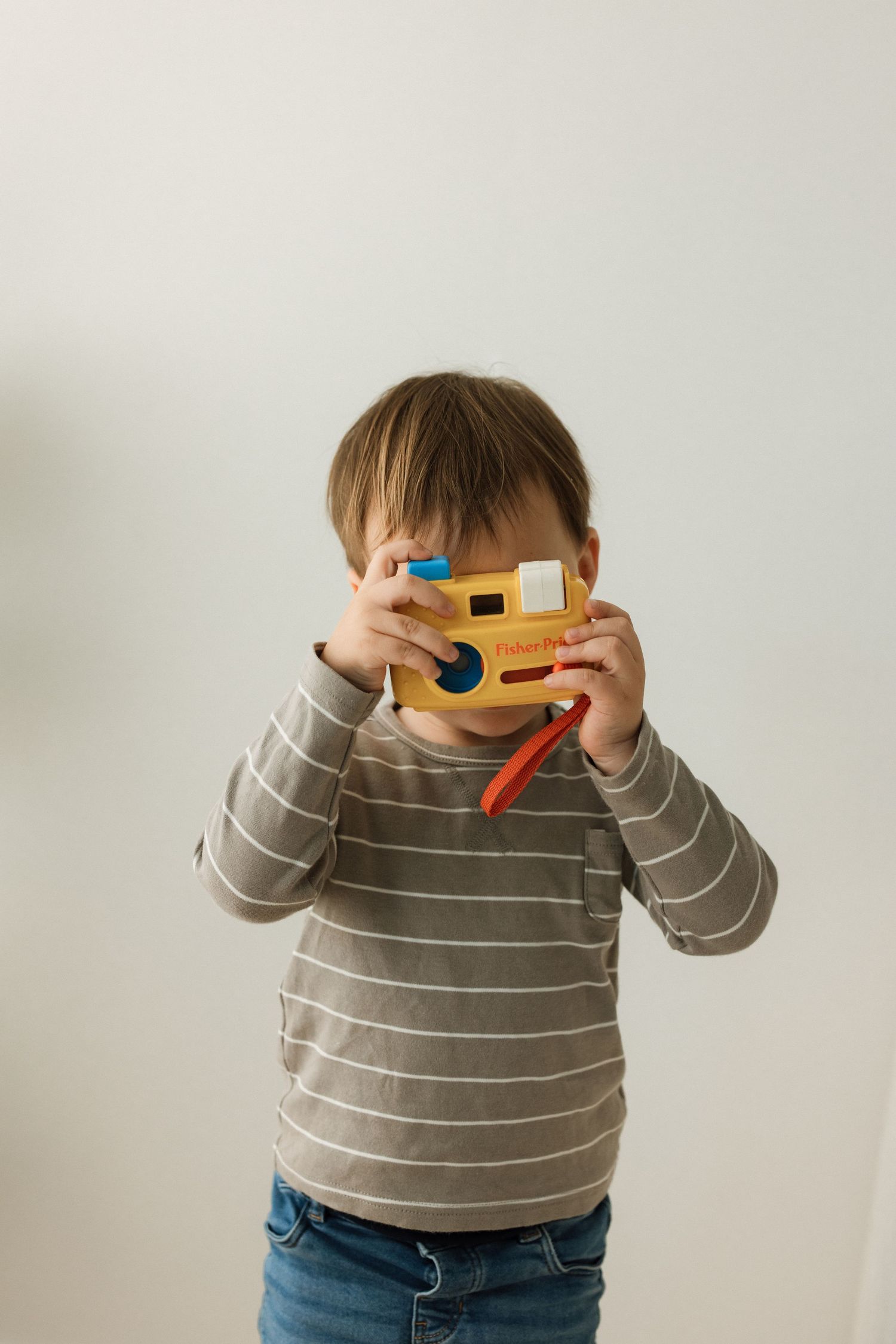 A small child in a striped sweater holds colorful 3D glasses up to their face against a white wall.