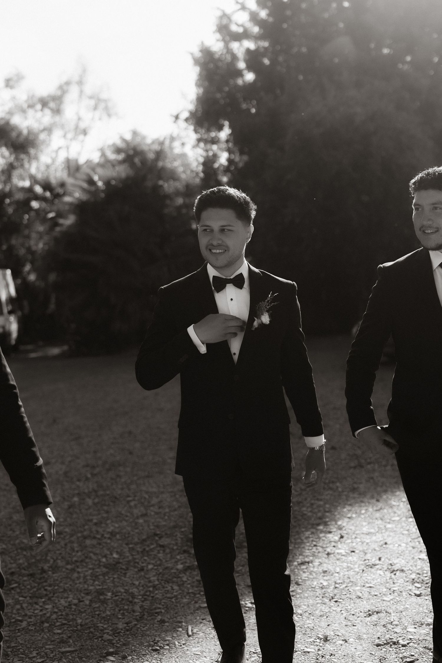 Groomsmen in black tuxedos walk together in dramatic backlit sunset setting.