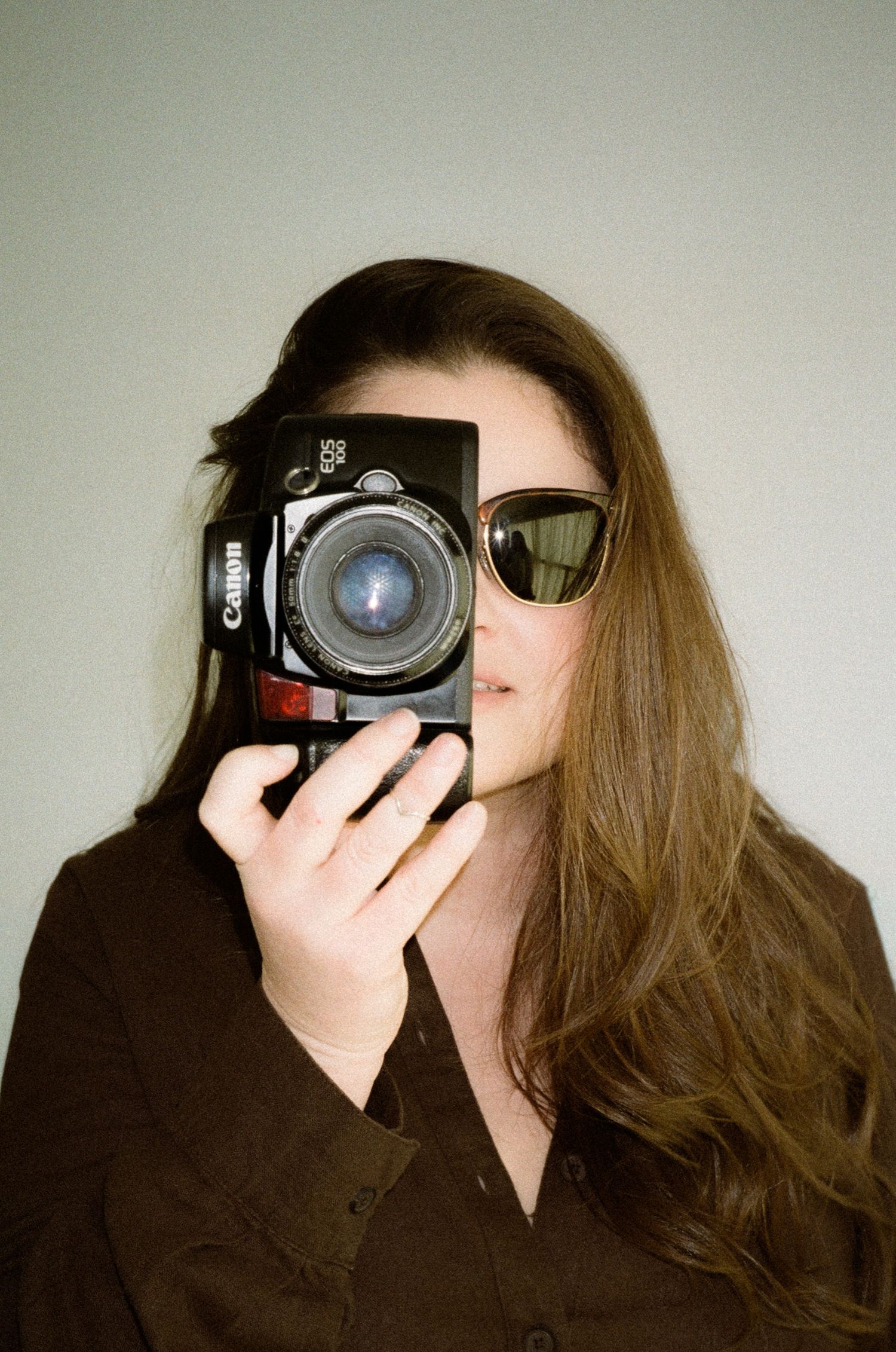 Woman holding a Canon film camera up to her face, wearing sunglasses and a dark shirt, photographed on film.