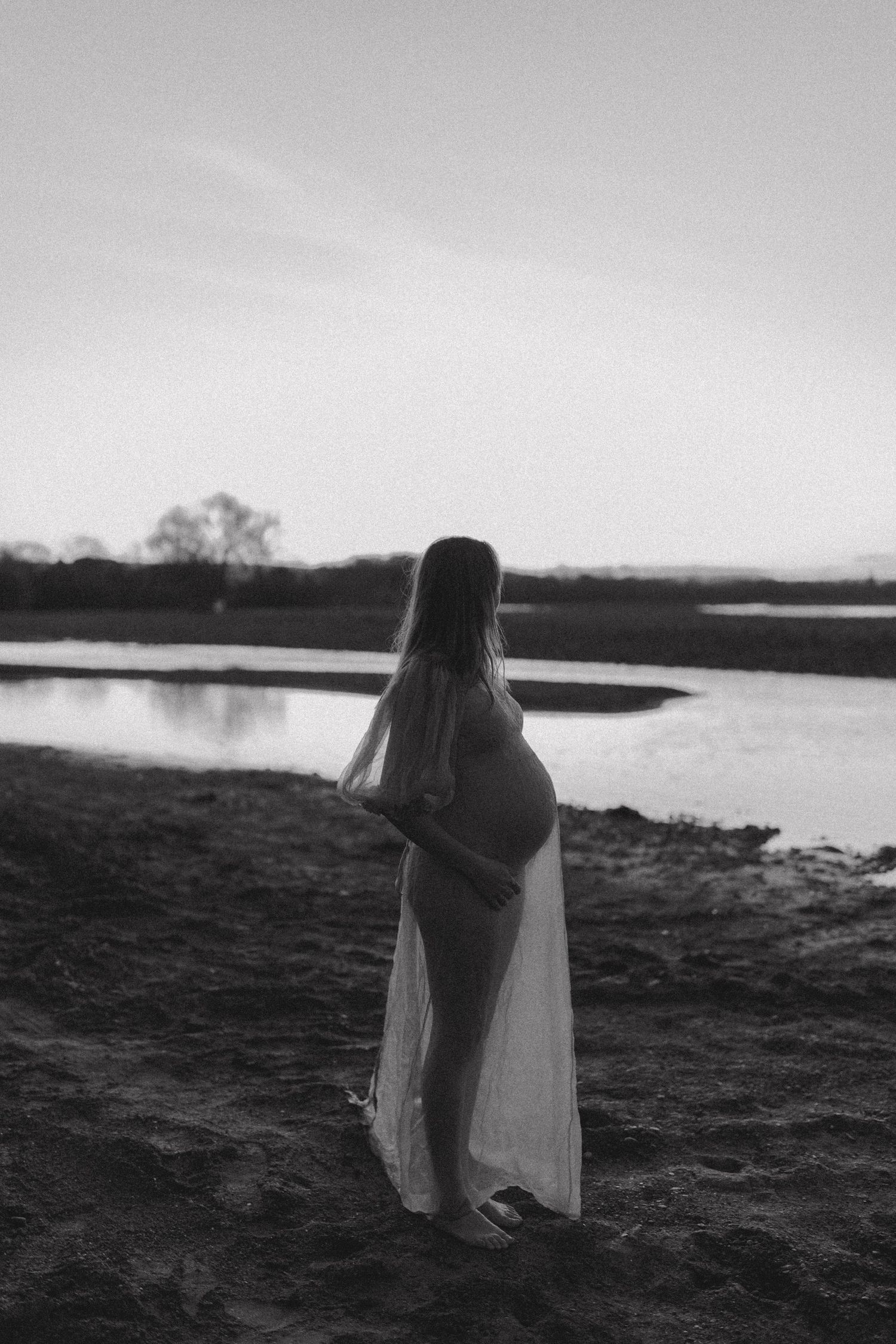 A silhouetted maternity figure standing by a lake at dusk in black and white.