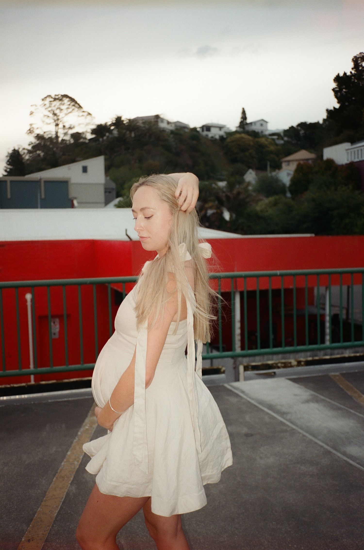 Pregnant woman in white dress poses on rooftop parking lot with red railing and green hills in background.
