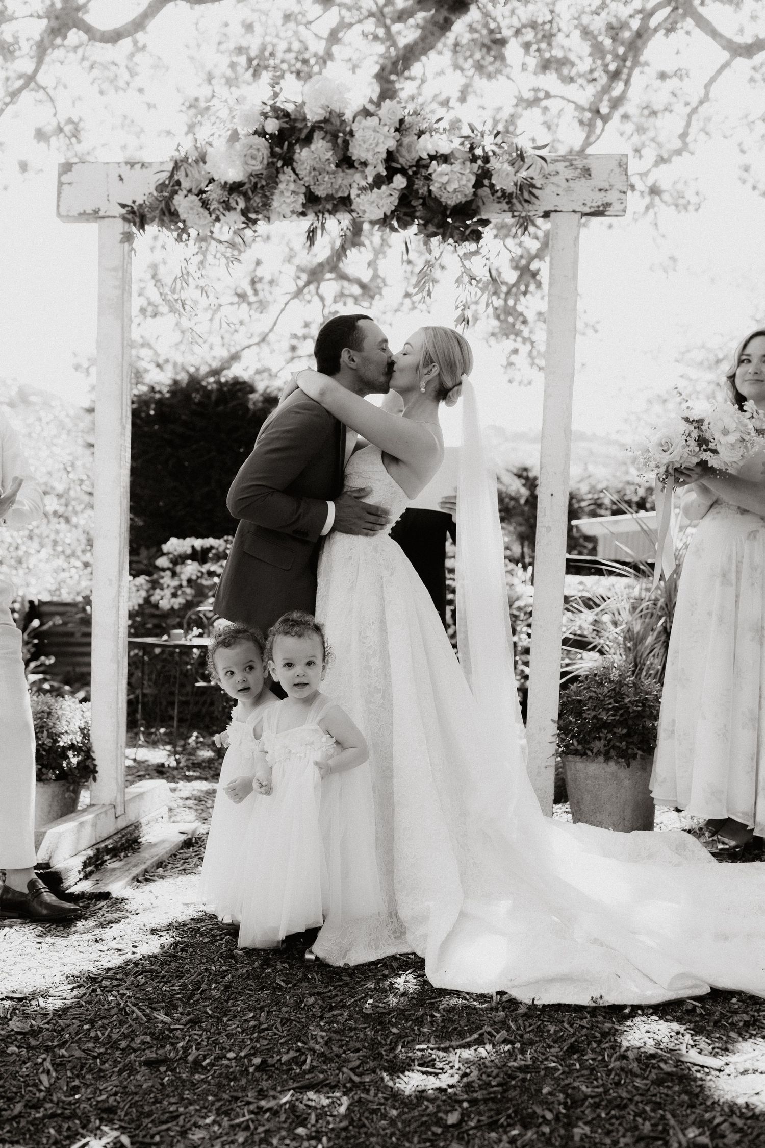 Black and white wedding photo of couple kissing under floral arch with flower girl standing in front.