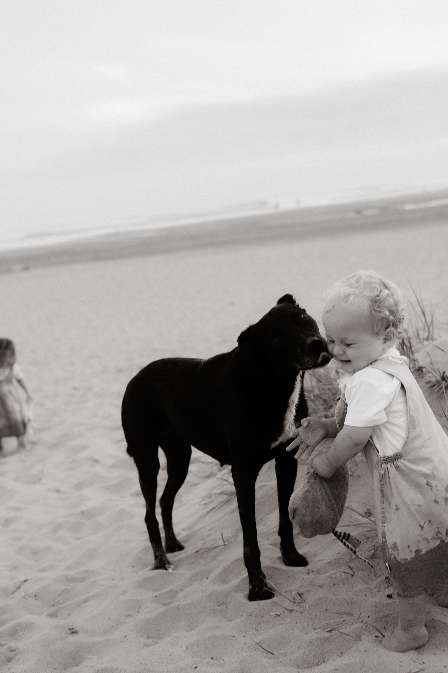 A small child and black dog share a tender moment together on a sandy beach in black and white photography.