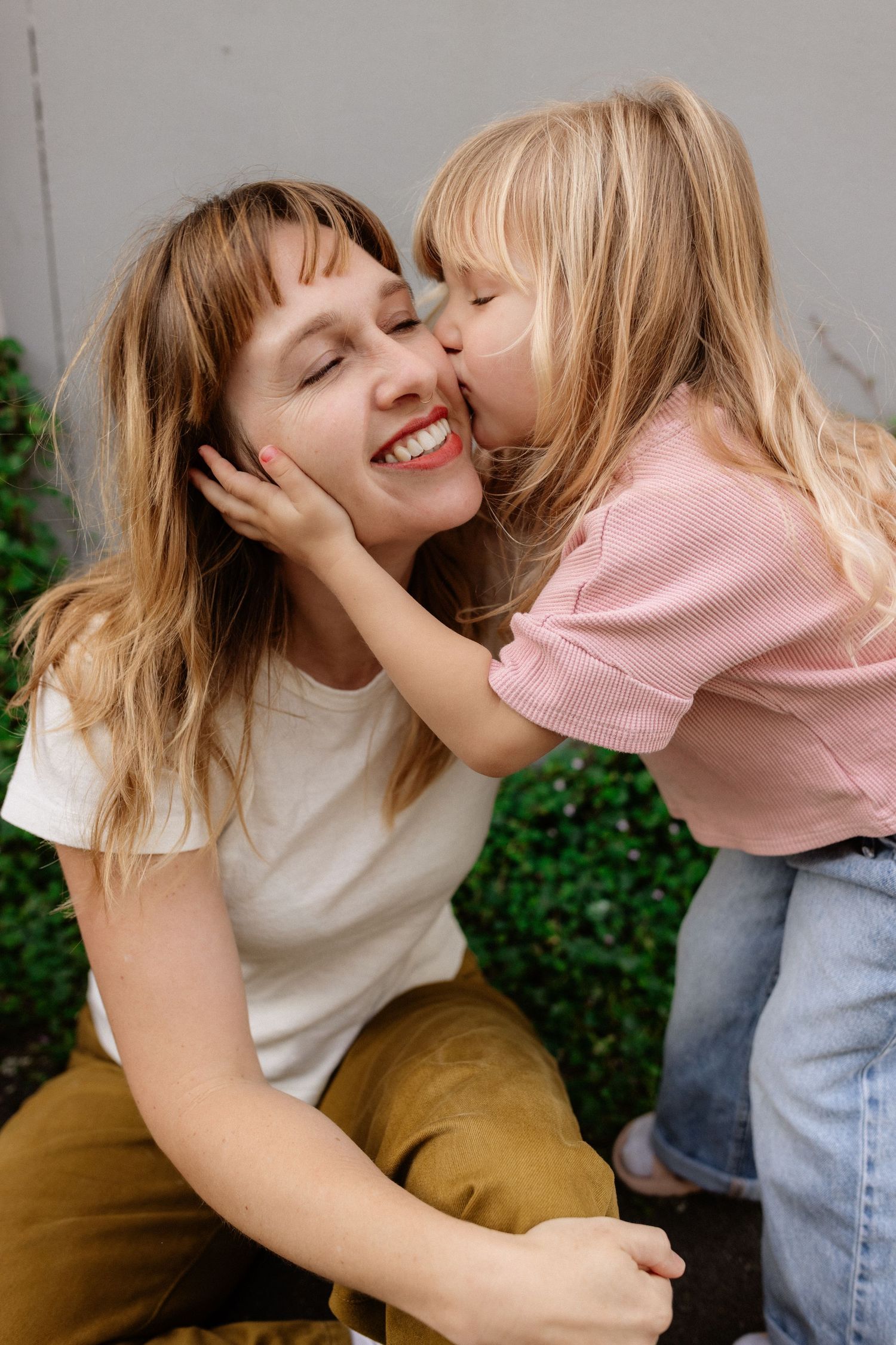 A young girl kissing her smiling mother on the cheek outdoors near green bushes.