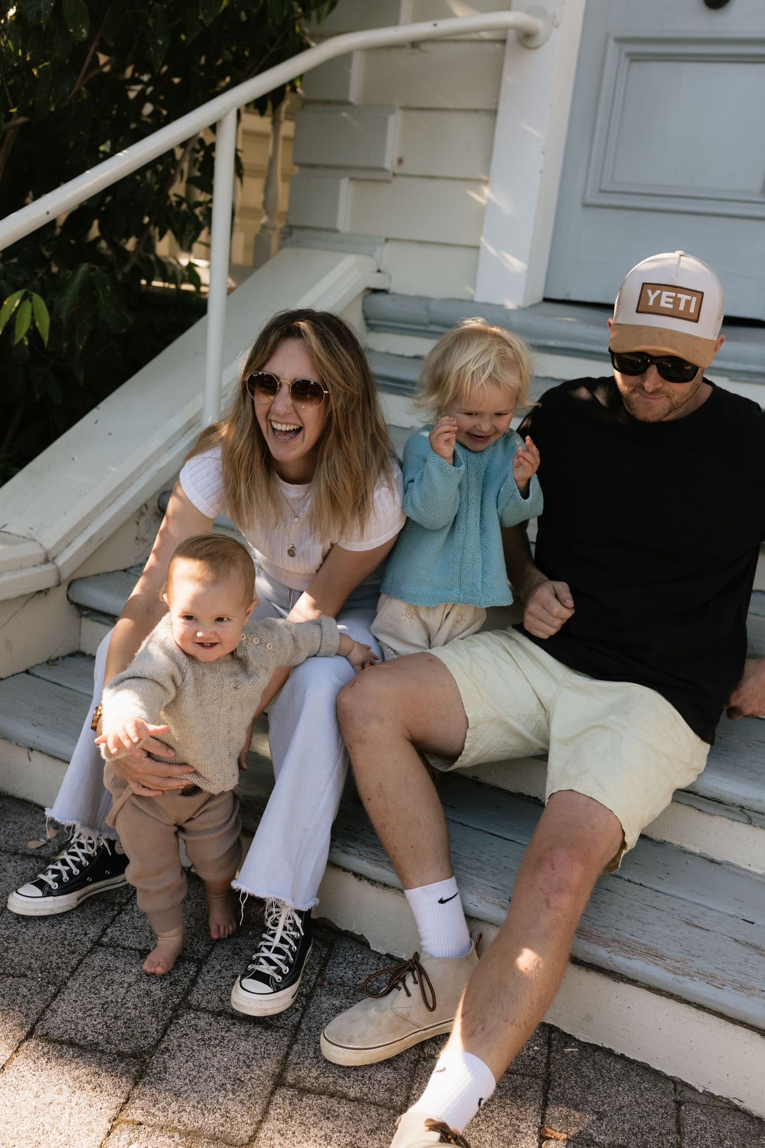 Happy family of four sitting on front porch steps, two parents laughing with two young toddlers on a sunny day.
