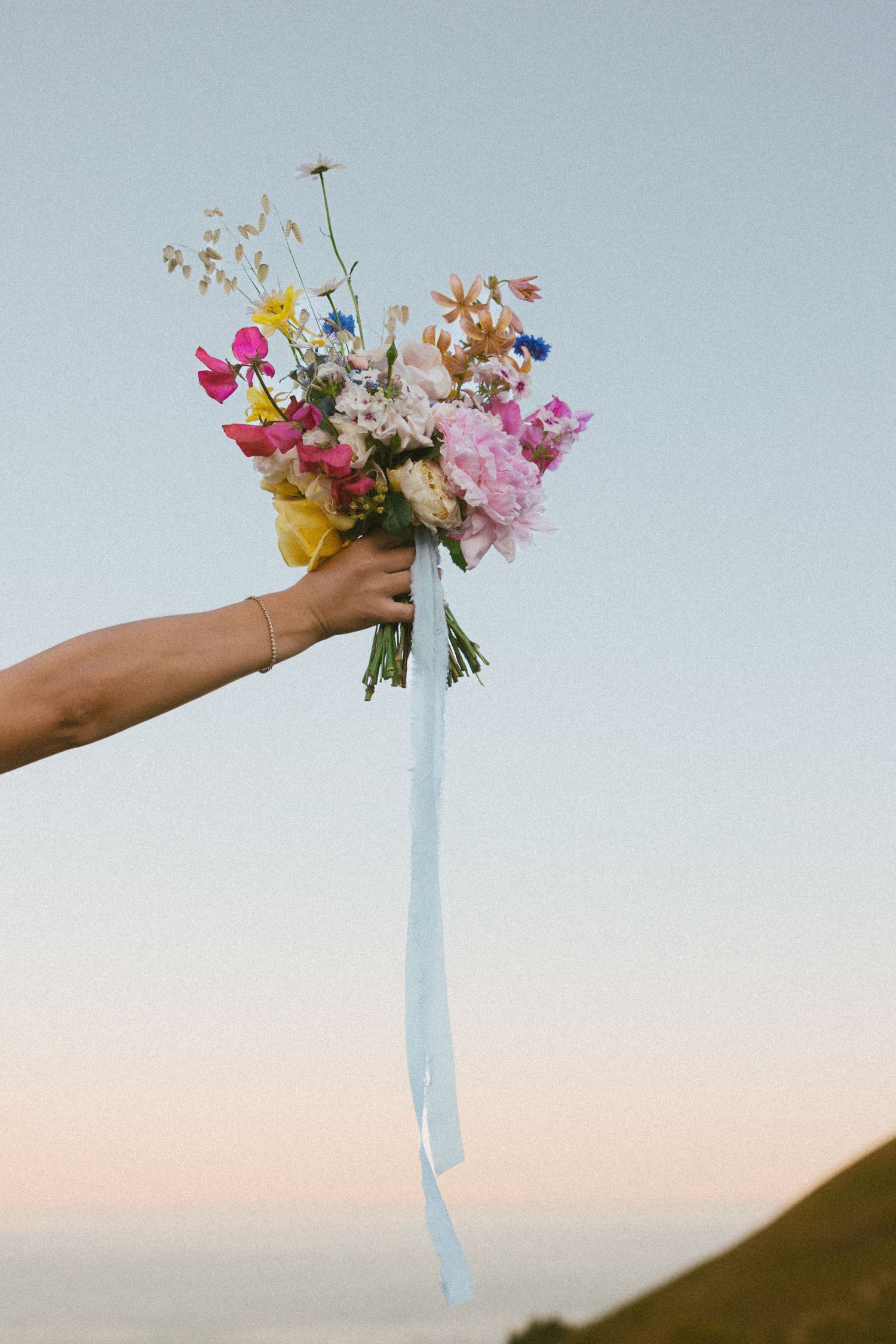 A hand holds a delicate bouquet of colorful flowers against a soft sunset sky with a light blue ribbon.