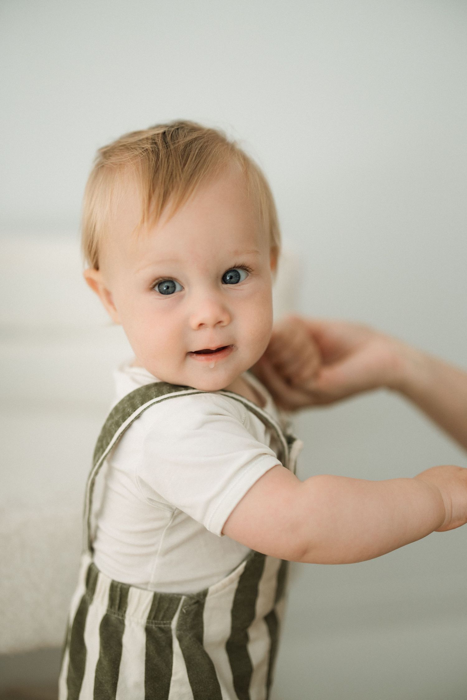 Baby wearing olive striped overalls and white shirt smiles while standing against light background.