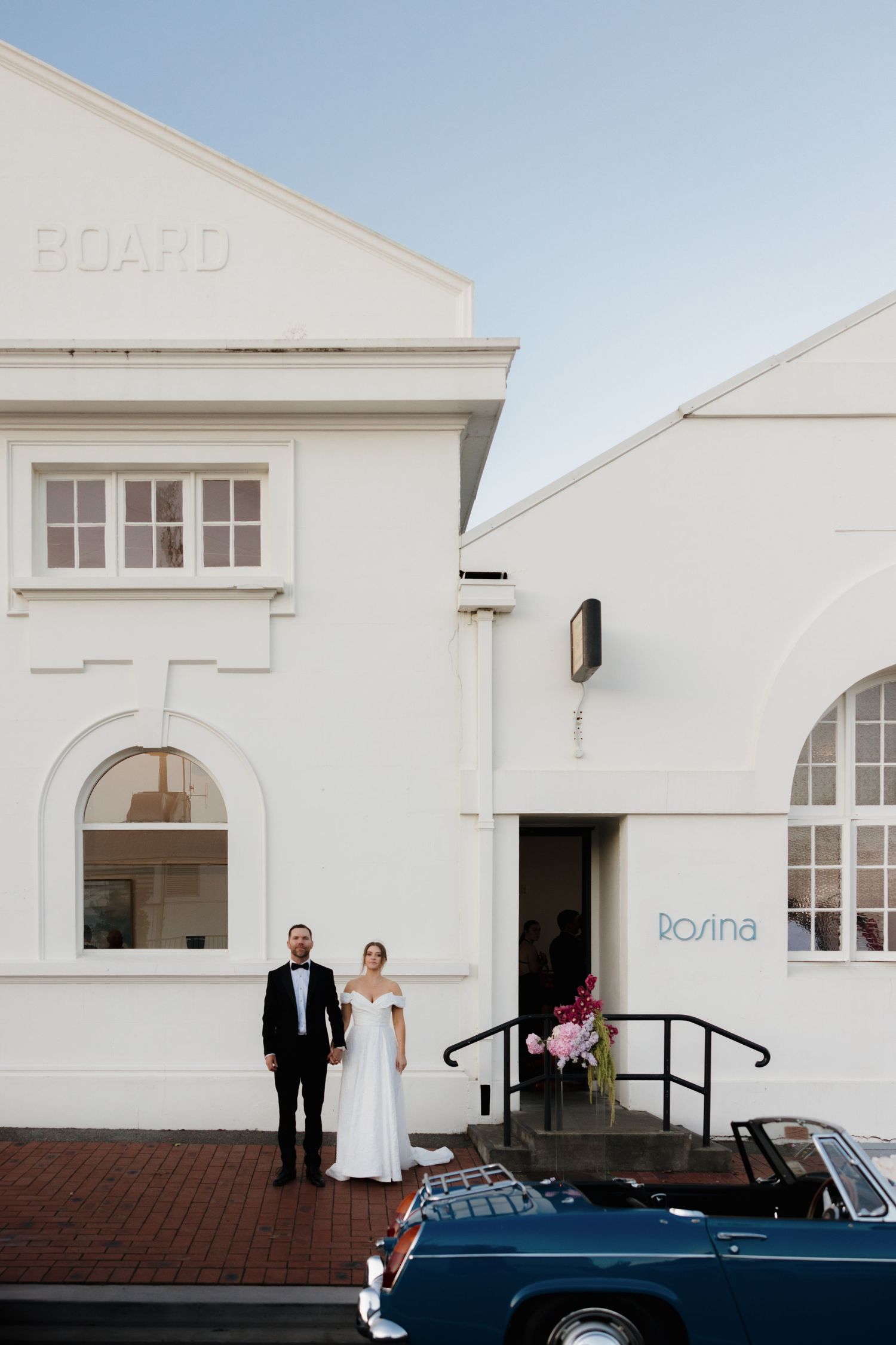 A newlywed couple stands outside a white building next to a classic blue car.