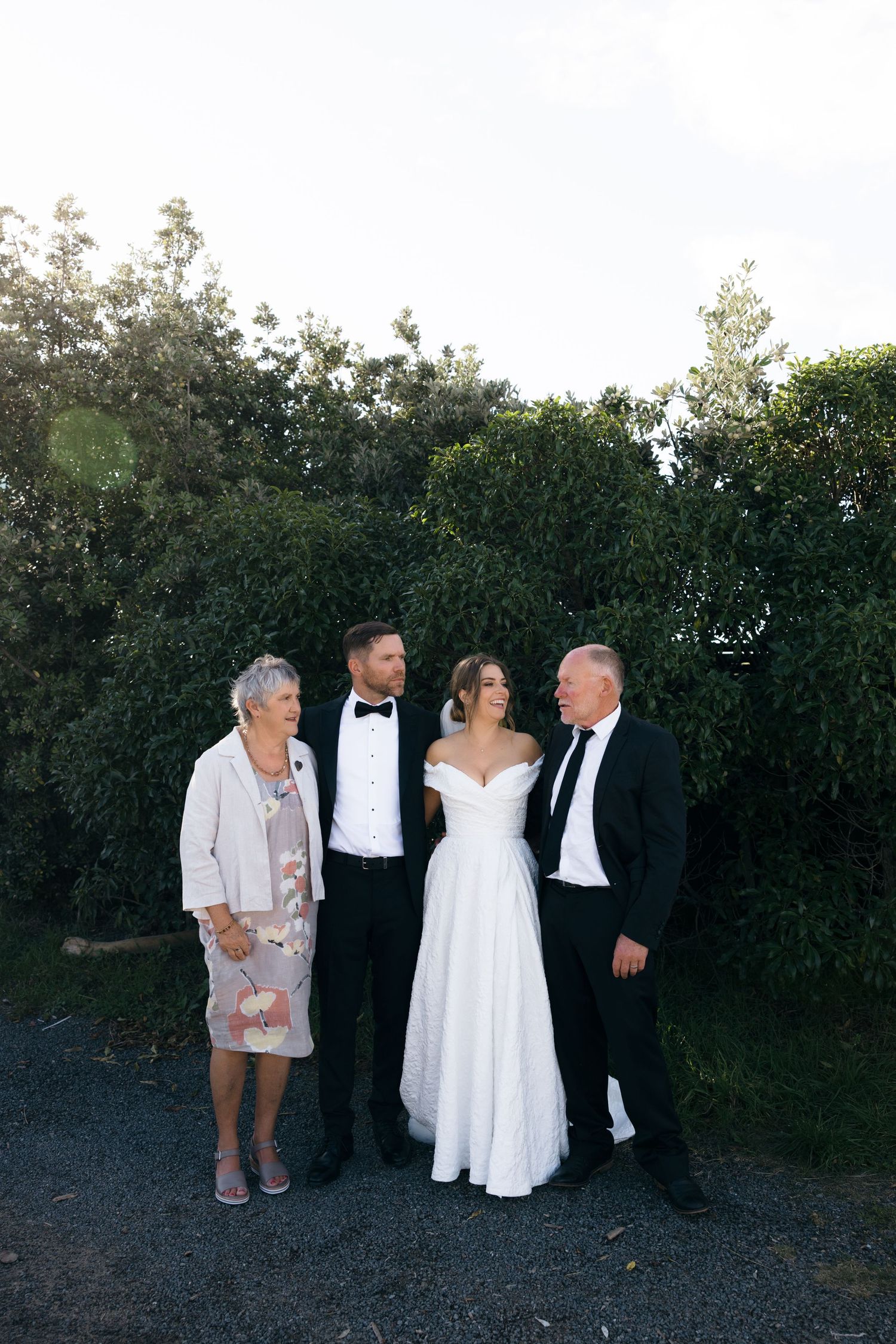 Wedding party poses casually against green hedge backdrop during outdoor celebration.