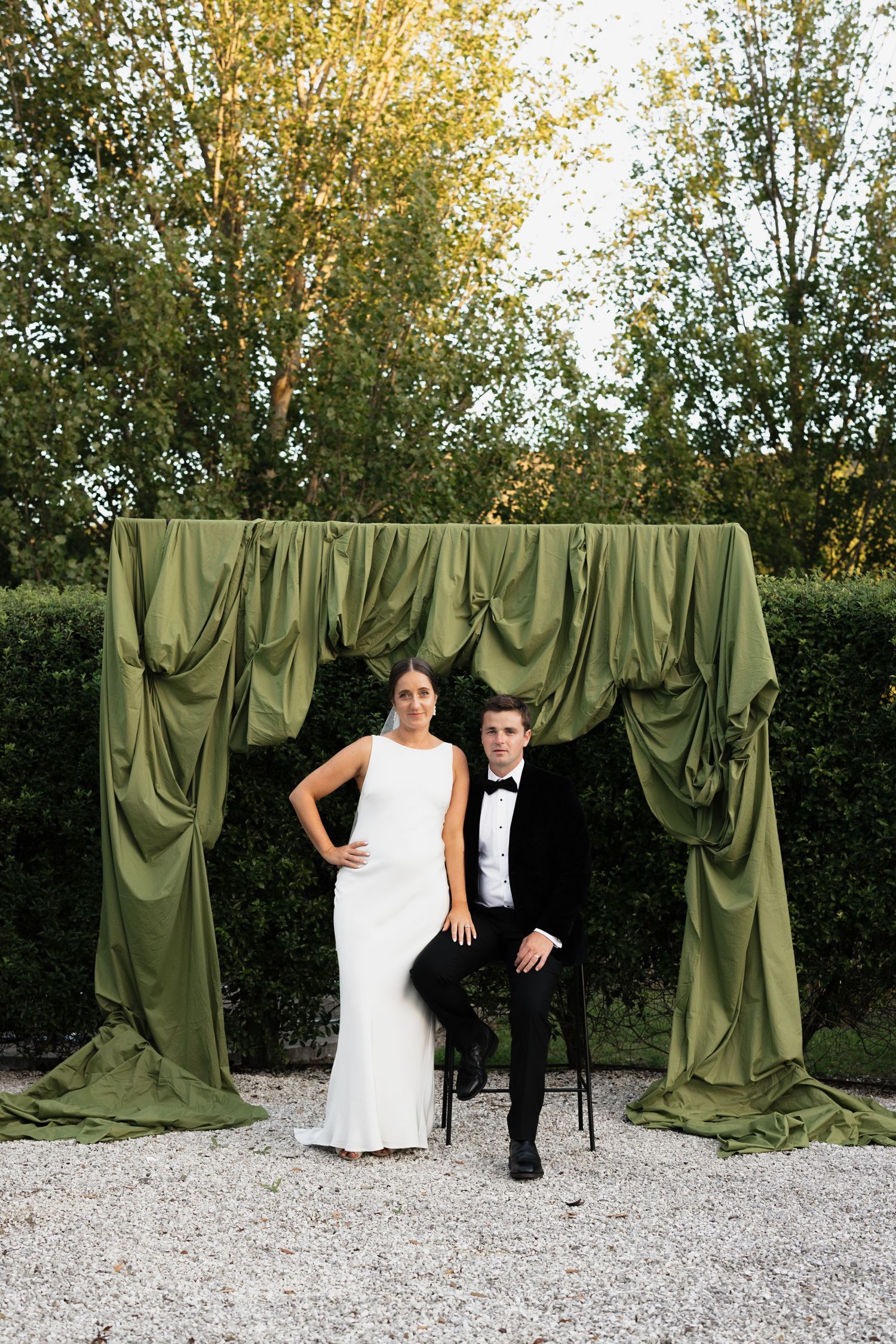 Wedding photoshoot against olive green draped backdrop on gravel path.