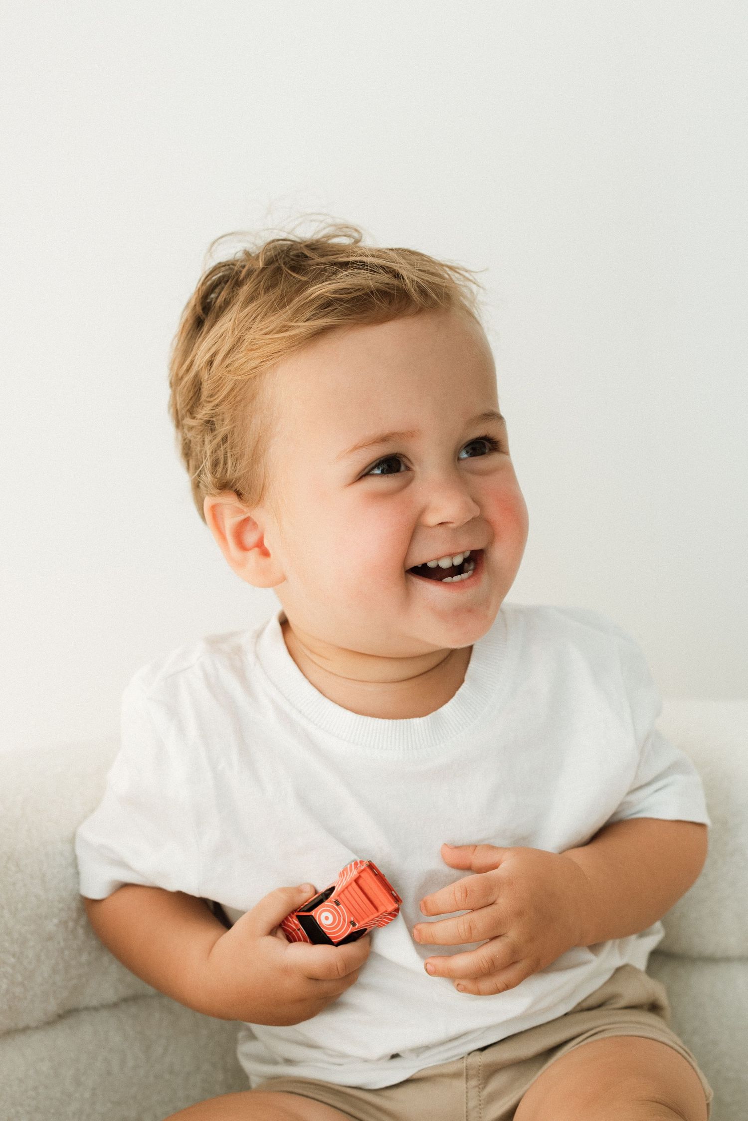 Laughing toddler in white t-shirt holding red toy on gray sofa.