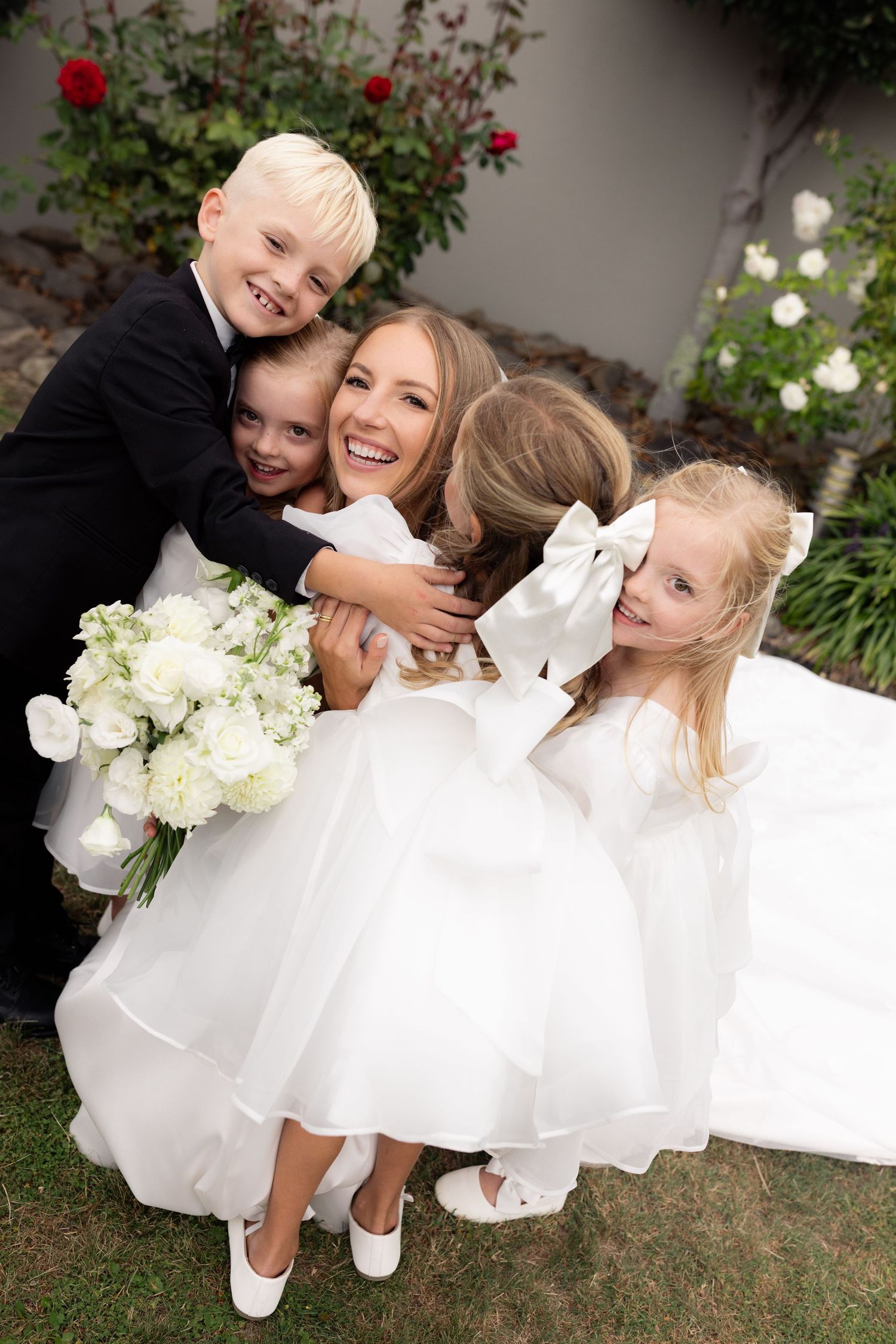 Two flower girls in white dresses twirl and play together in a garden setting with blooming flowers.
