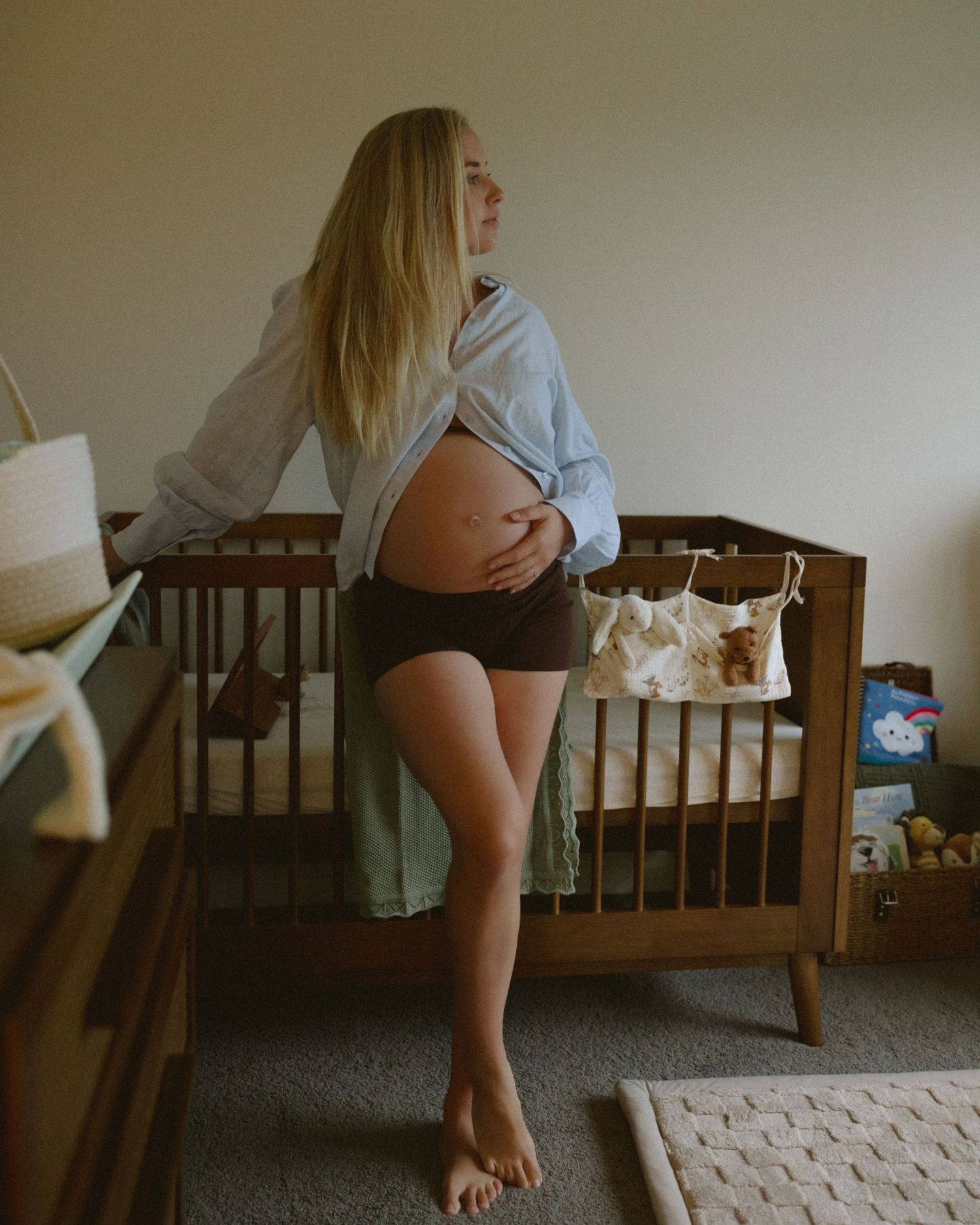 Pregnant woman in blue shirt cradling baby bump while standing in a nursery with wooden cribs.