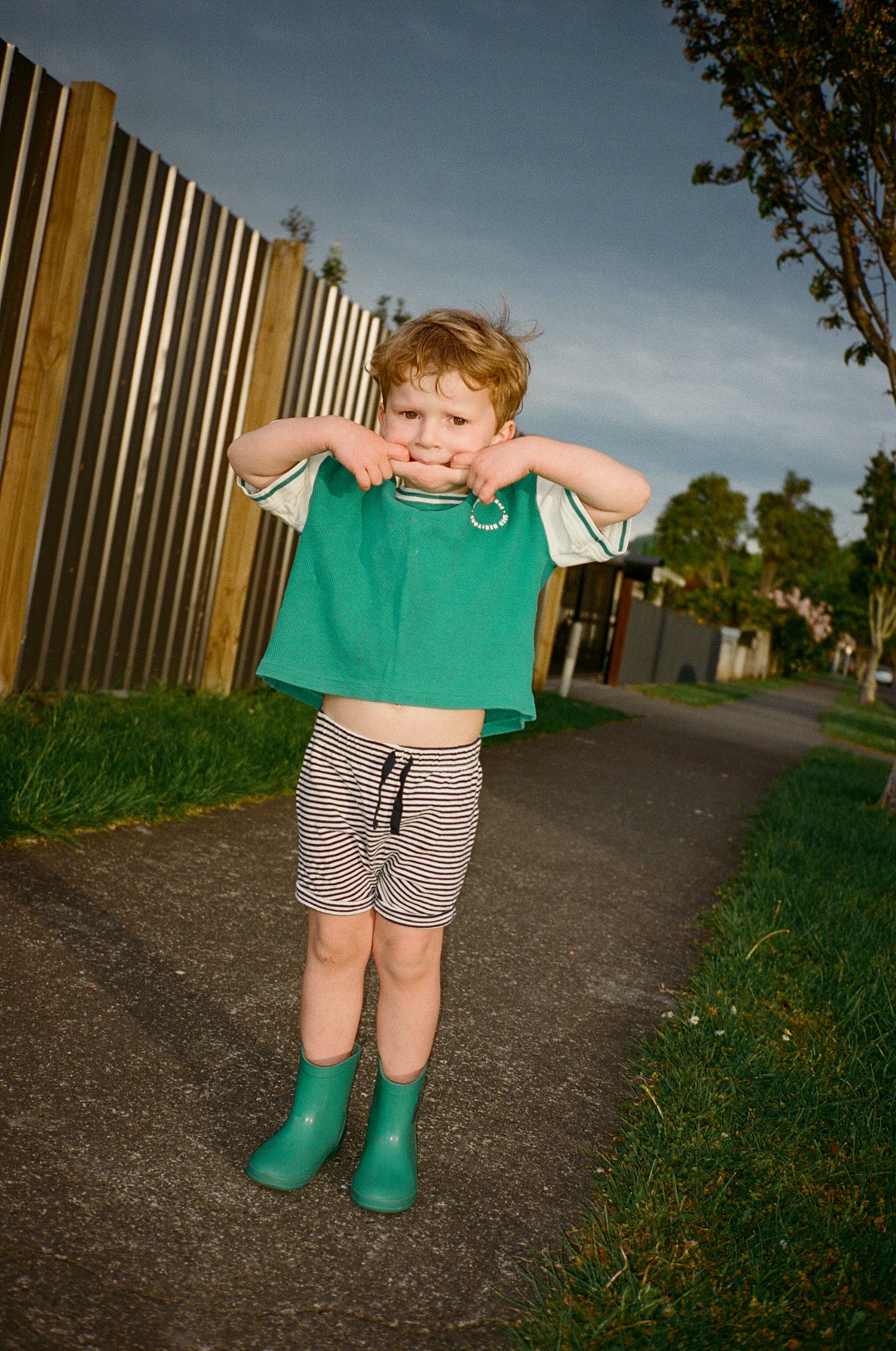Small child wearing green rain boots and striped shorts plays on a path next to a wooden fence.