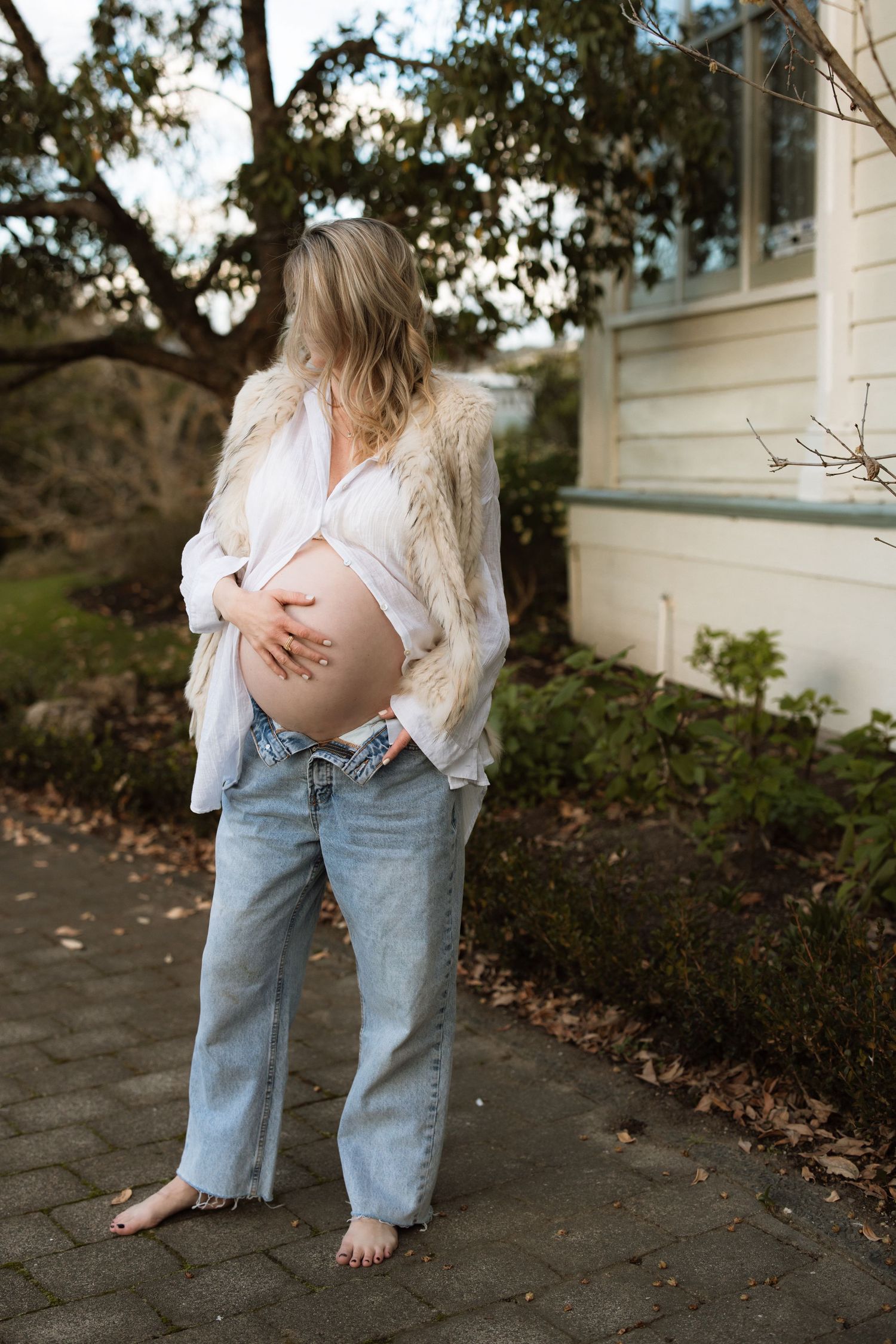 A pregnant woman in a white lace top and loose jeans stands barefoot against a white building exterior.