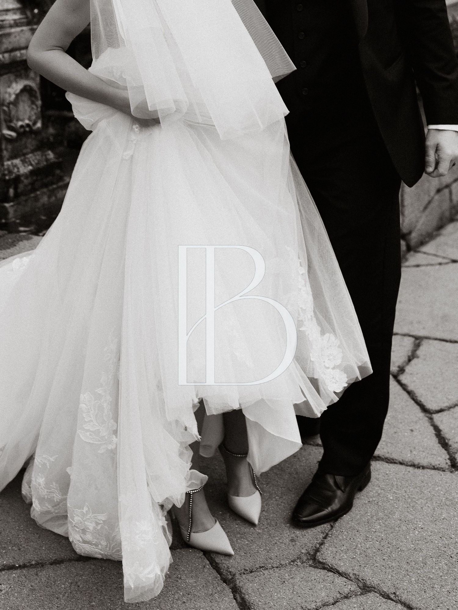 Black and white photo of a bride in a tulle wedding gown with lace appliqués and pointed heels standing beside a groom in a dark suit.