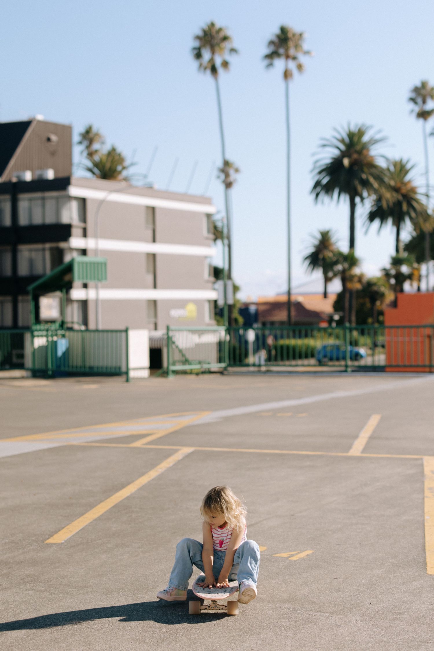 Person lying on empty parking lot with palm trees and buildings in background.