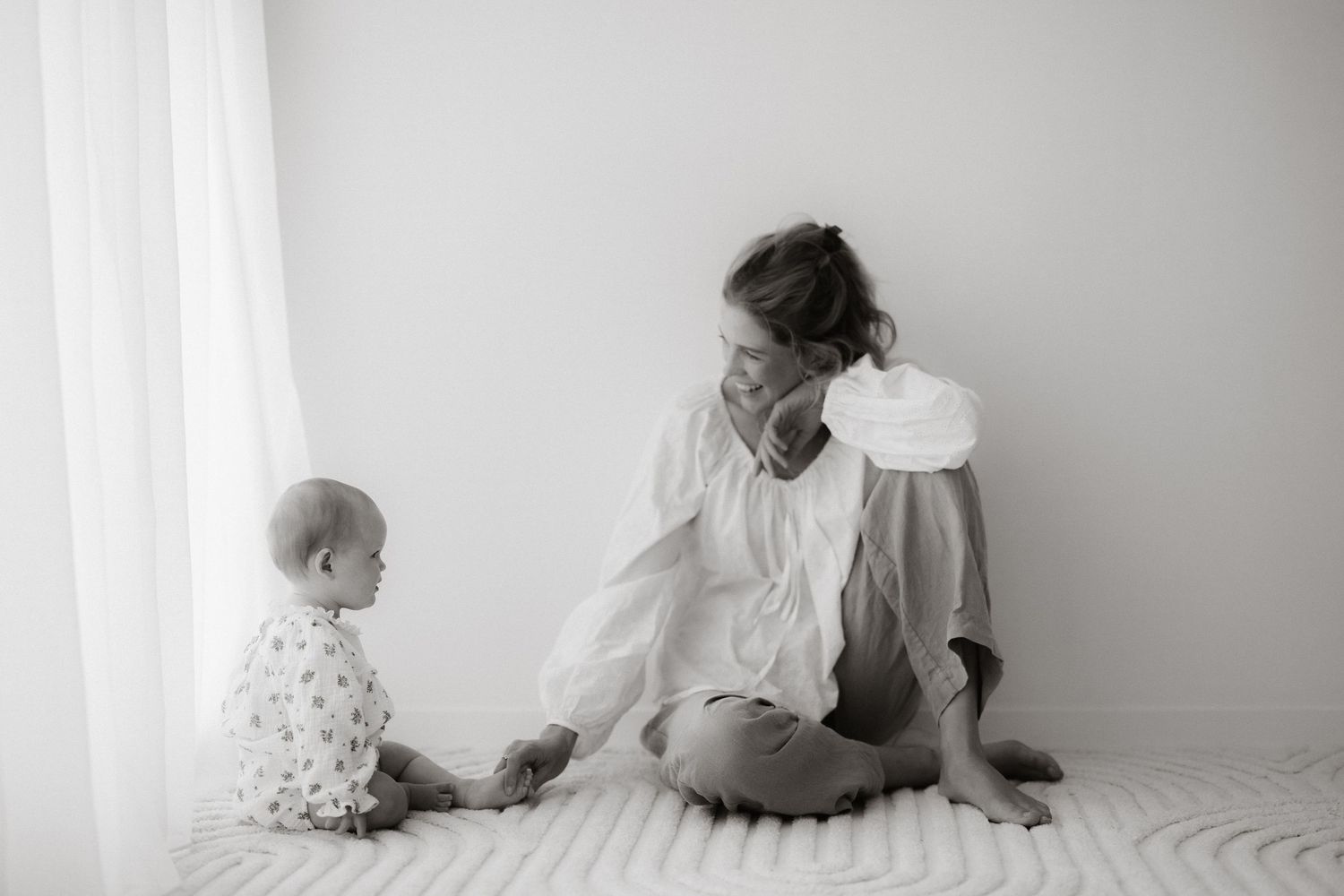 Black and white photo of an intimate parent-child moment sitting together on a bed near a window.