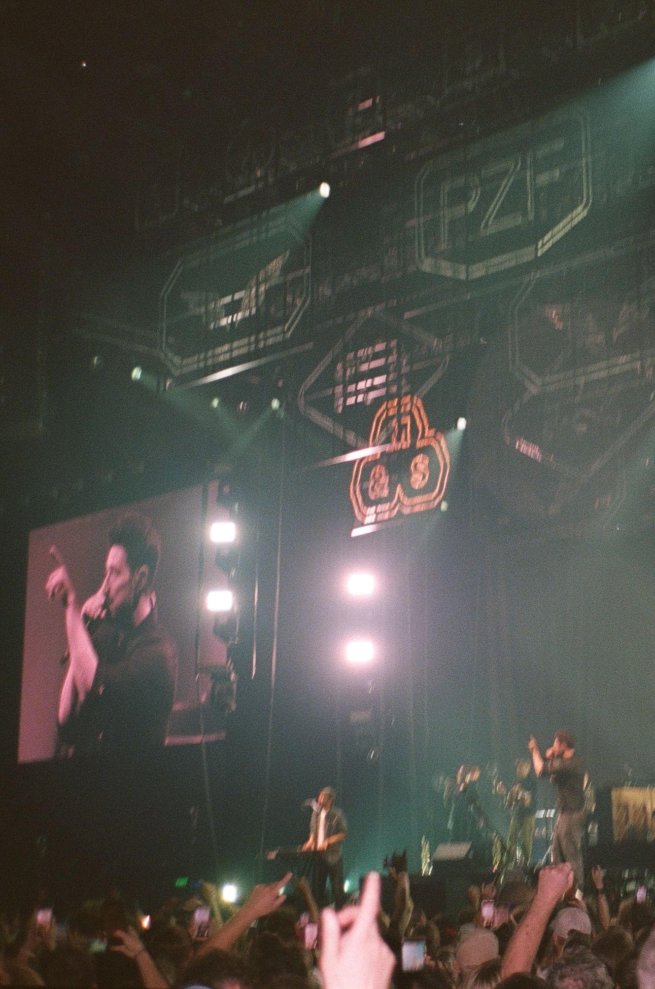 Energetic concert crowd with bright stage lights, large LED screen, and neon logo display at a live music event.