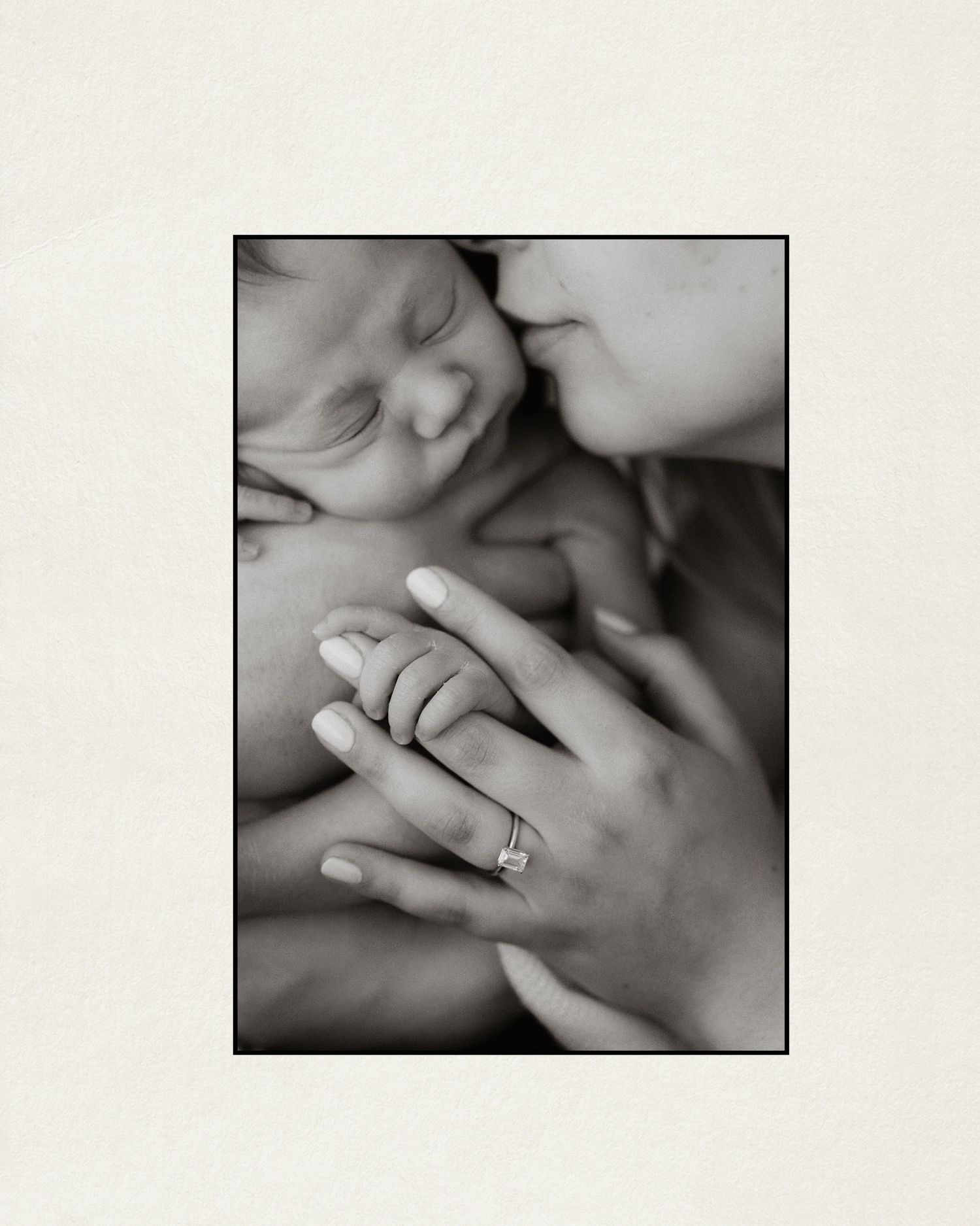 Black and white photo of a mother kissing her newborn baby while holding tiny hands, wearing a diamond engagement ring.