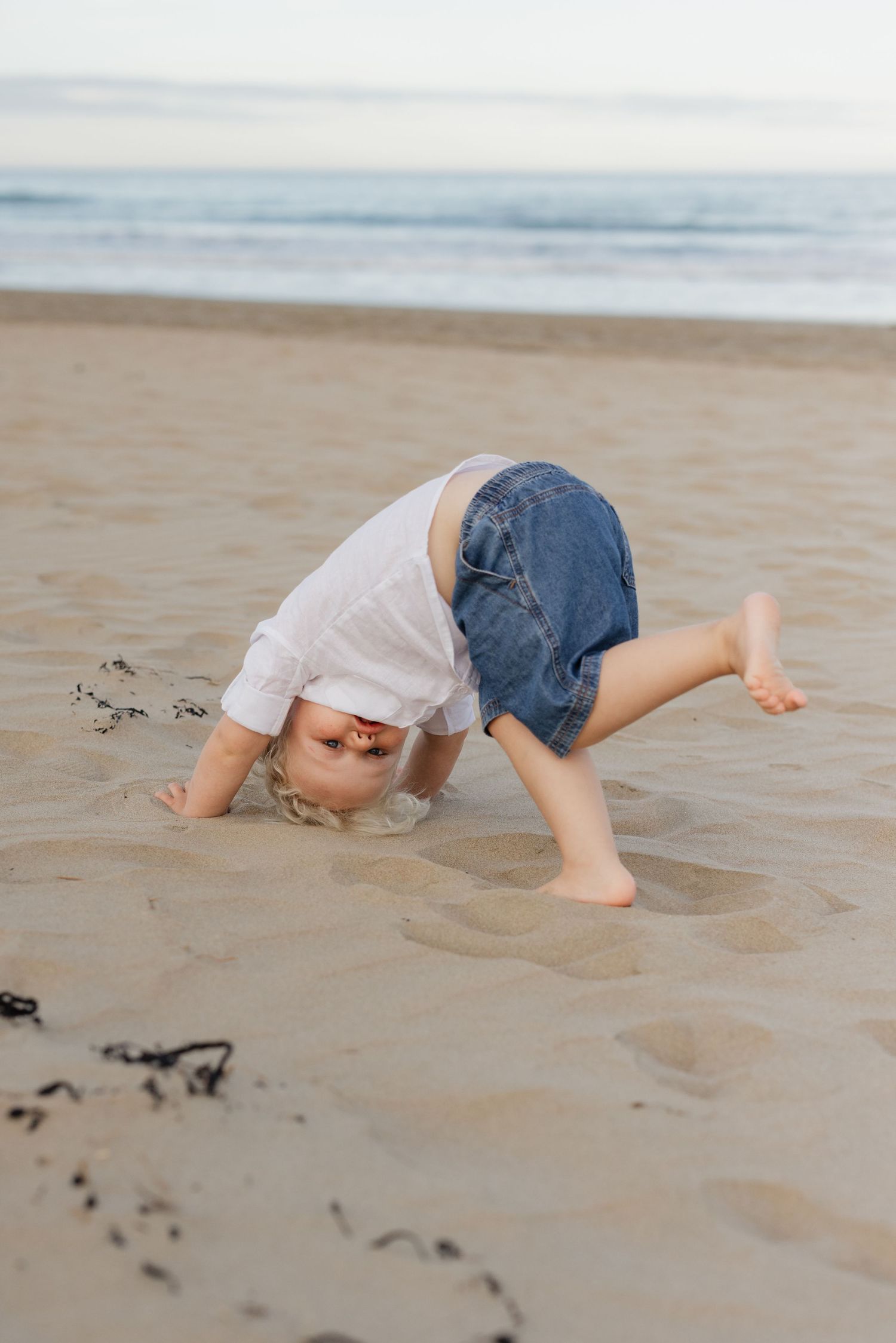 A child in denim shorts and white shirt bends over playfully on a sandy beach near the ocean.