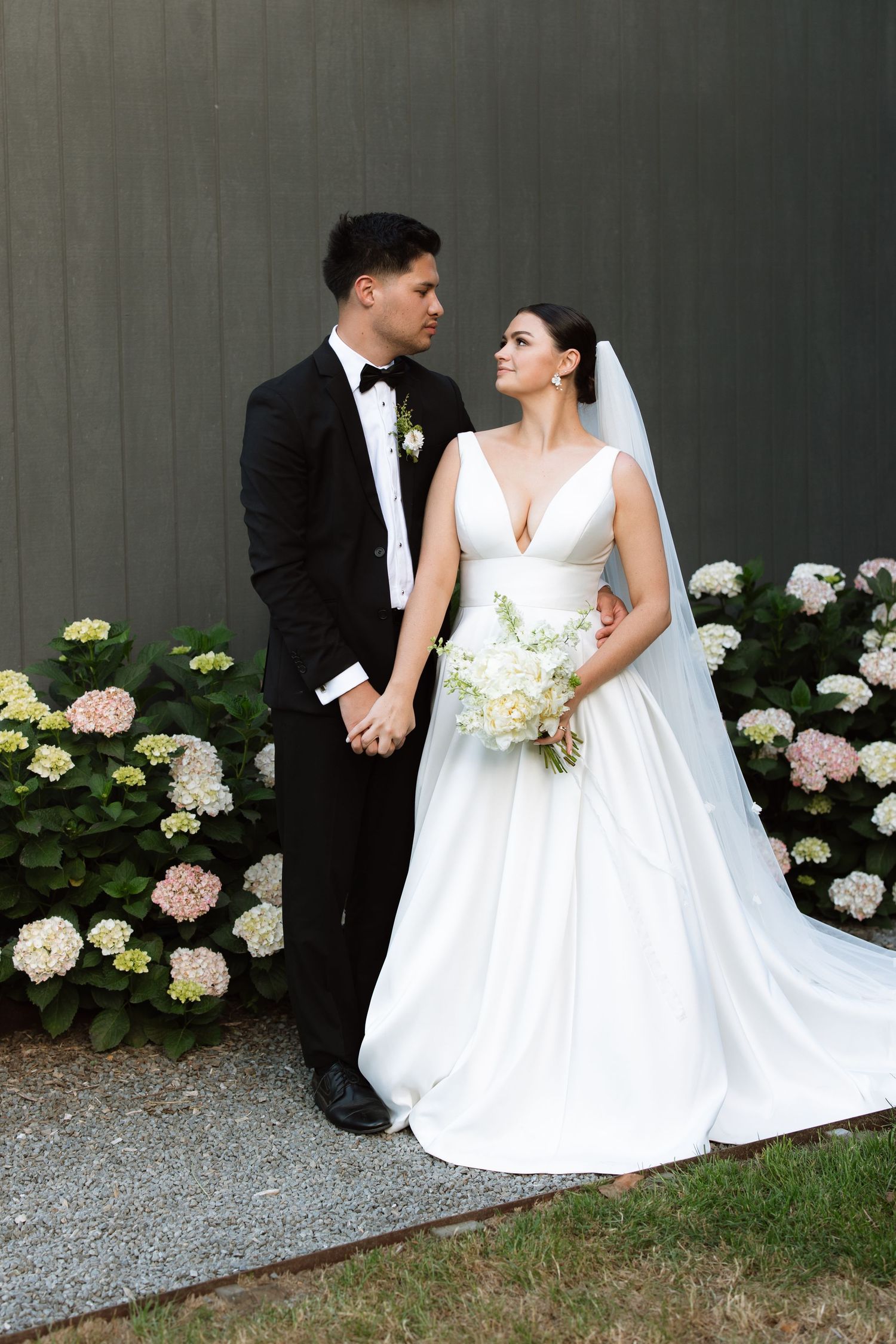 Wedding couple poses against grey wall with hydrangea flowers in elegant formal attire.