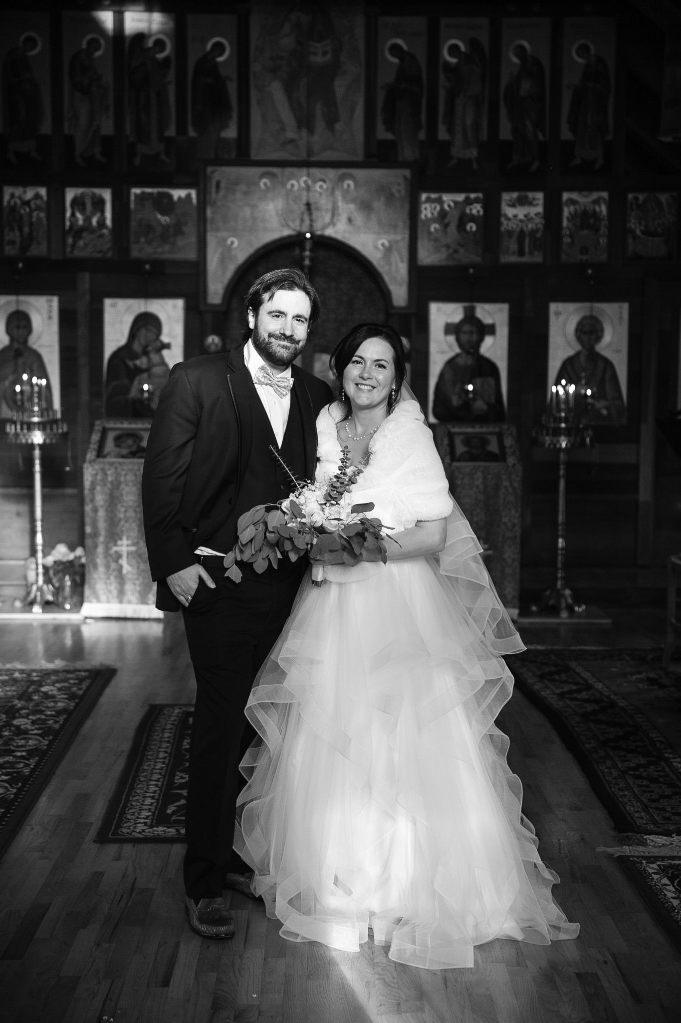 Black and white photo of a smiling bride and groom posing together inside an Orthodox church with religious icons.
