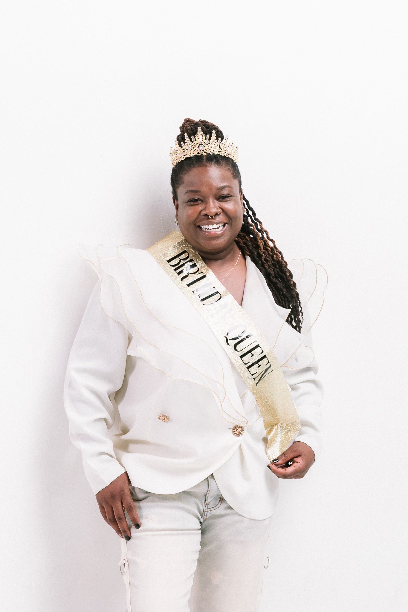Smiling woman in white blazer wearing a crown and Bride Queen sash posing against a white background.