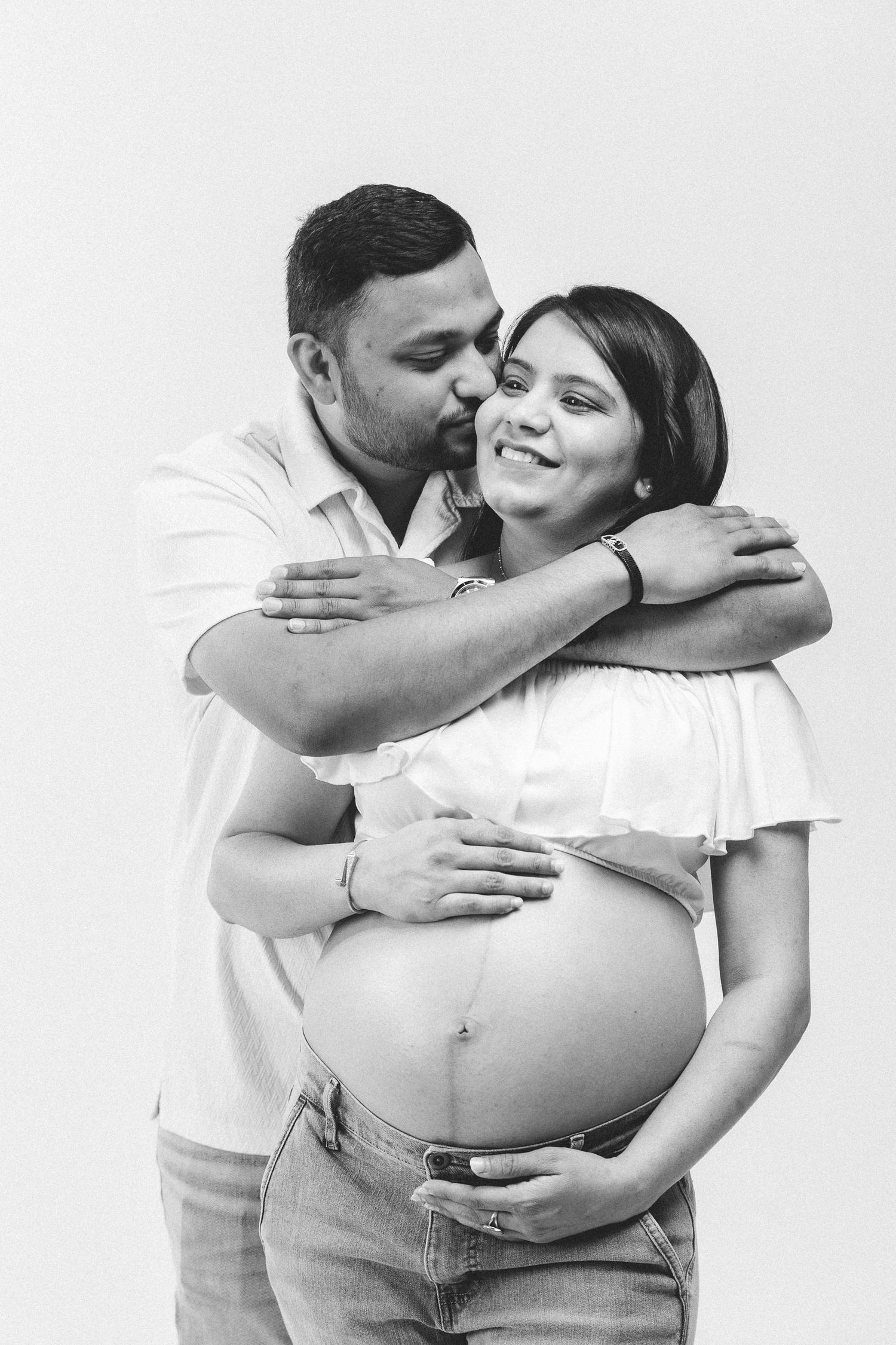 Black and white photo of a man kissing pregnant woman on cheek while embracing her baby bump together.
