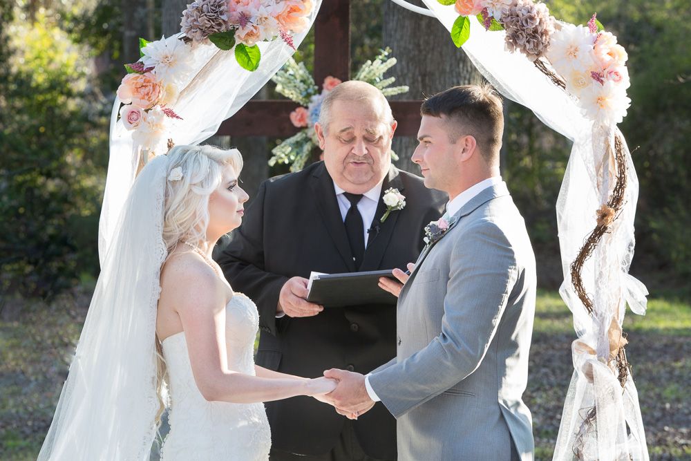 Bride and groom exchanging vows under a floral arch with an officiant at an outdoor wedding ceremony.