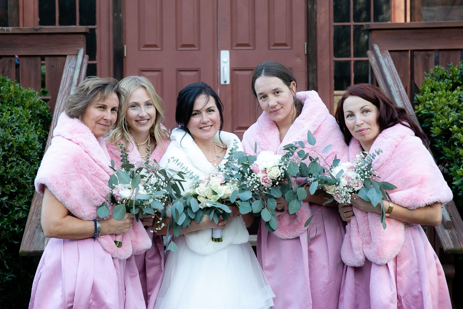 Bride in white gown with four bridesmaids in pink fur shawls holding floral bouquets posing outdoors near a wooden door.